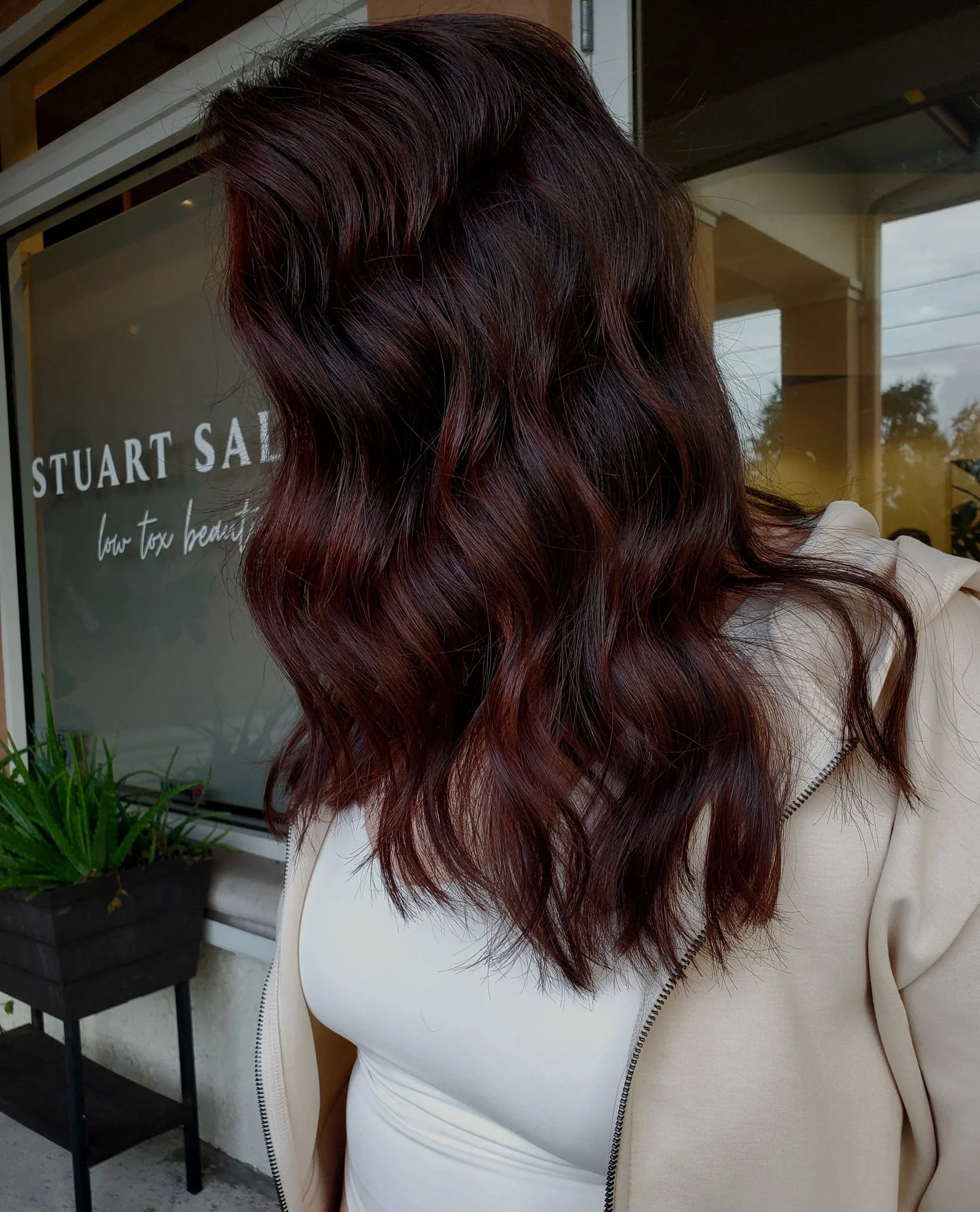 Woman with wavy, dark red hair standing outside in front of a storefront window with the words 'Stuart Sal' and 'low tax beauty' on it, wearing a light-colored jacket and tank top, next to a small black railing with a potted plant.