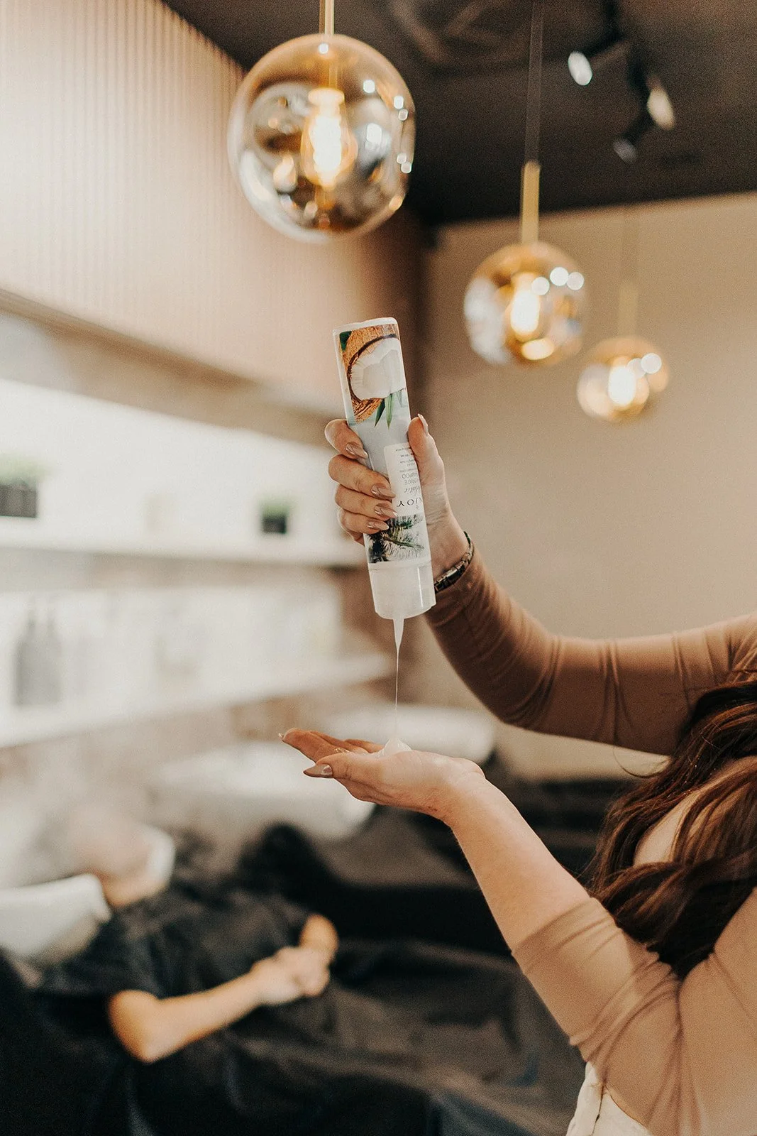 A woman is pouring lotion from a container with a tropical design into her hand, with a child lying on a couch in the background.