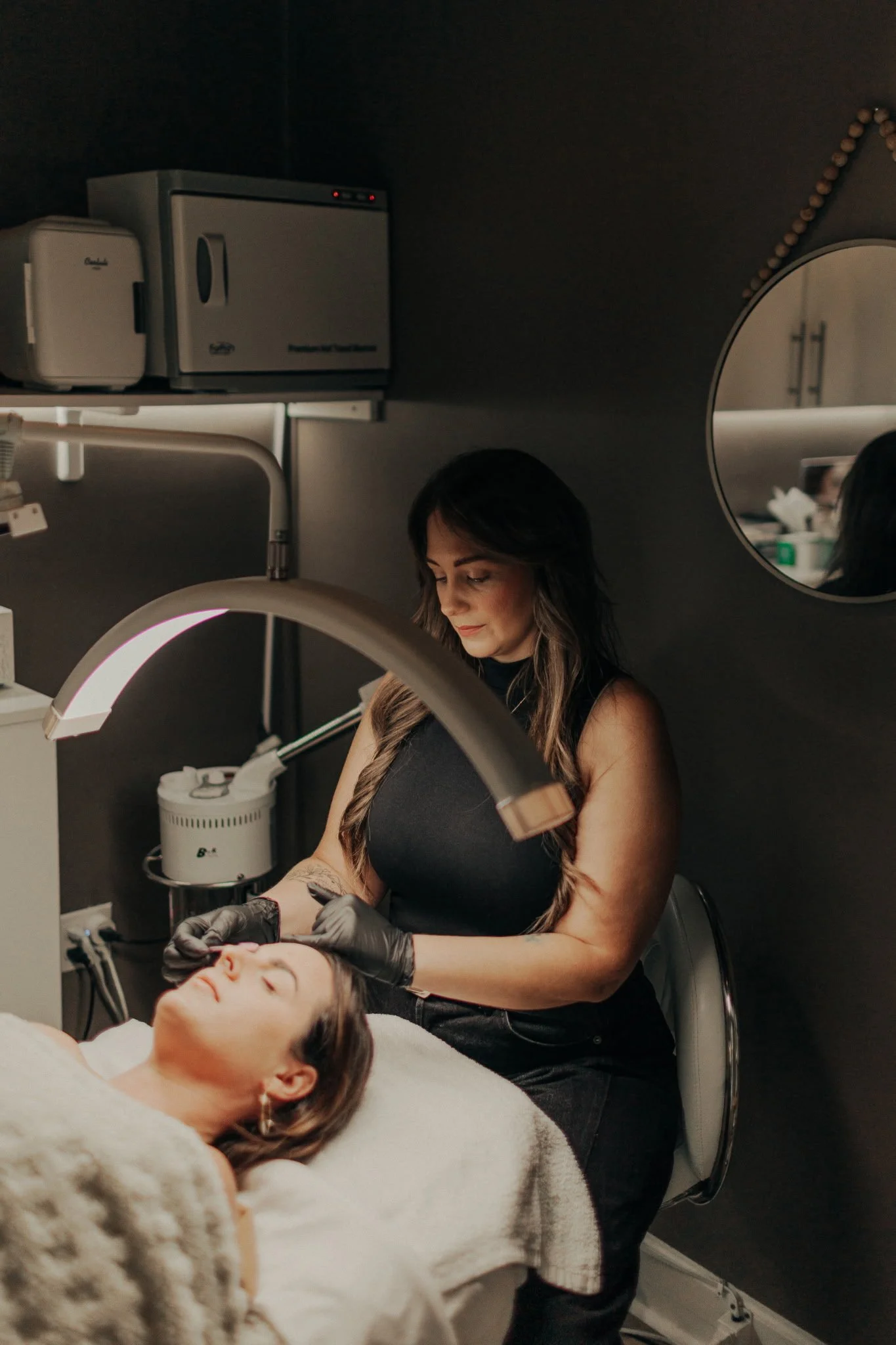 Cozy spa room with white robe hanging, massage table with blanket and pillow, overhead light, shelves with skincare products, and circular mirror.