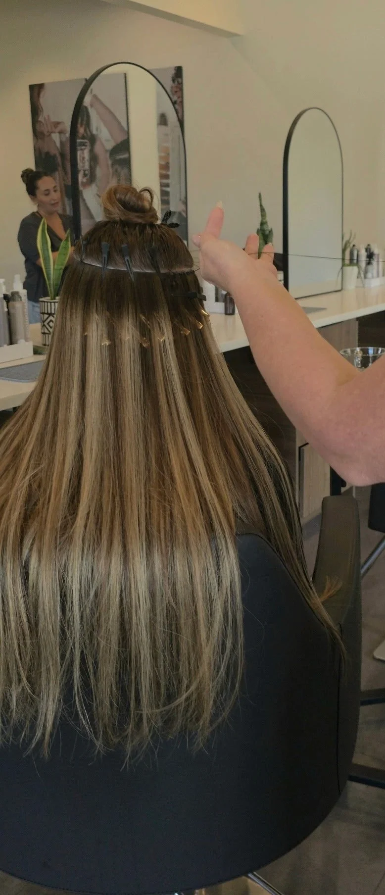 A woman with long blonde hair at a salon, getting her hair styled. A stylist is pointing and working on her hair, which is sectioned and clipped.