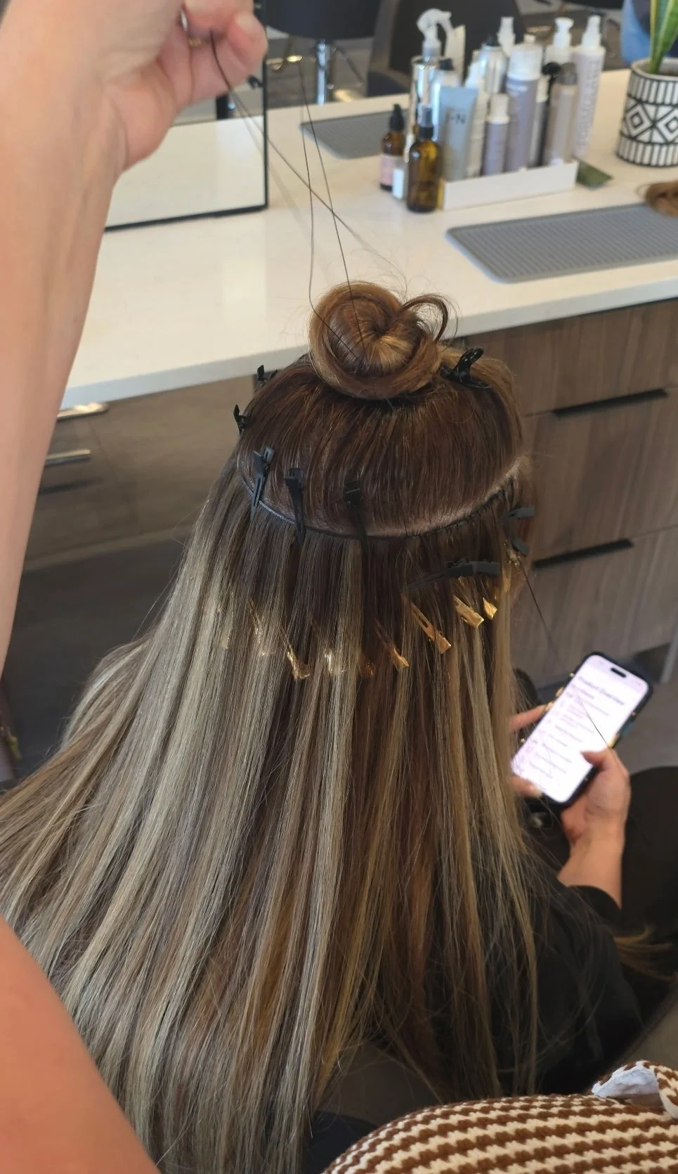 A woman with long blonde hair sits at a hair salon, clamped with clips, while a stylist uses a needle to weave hair extensions. She is looking at her smartphone.