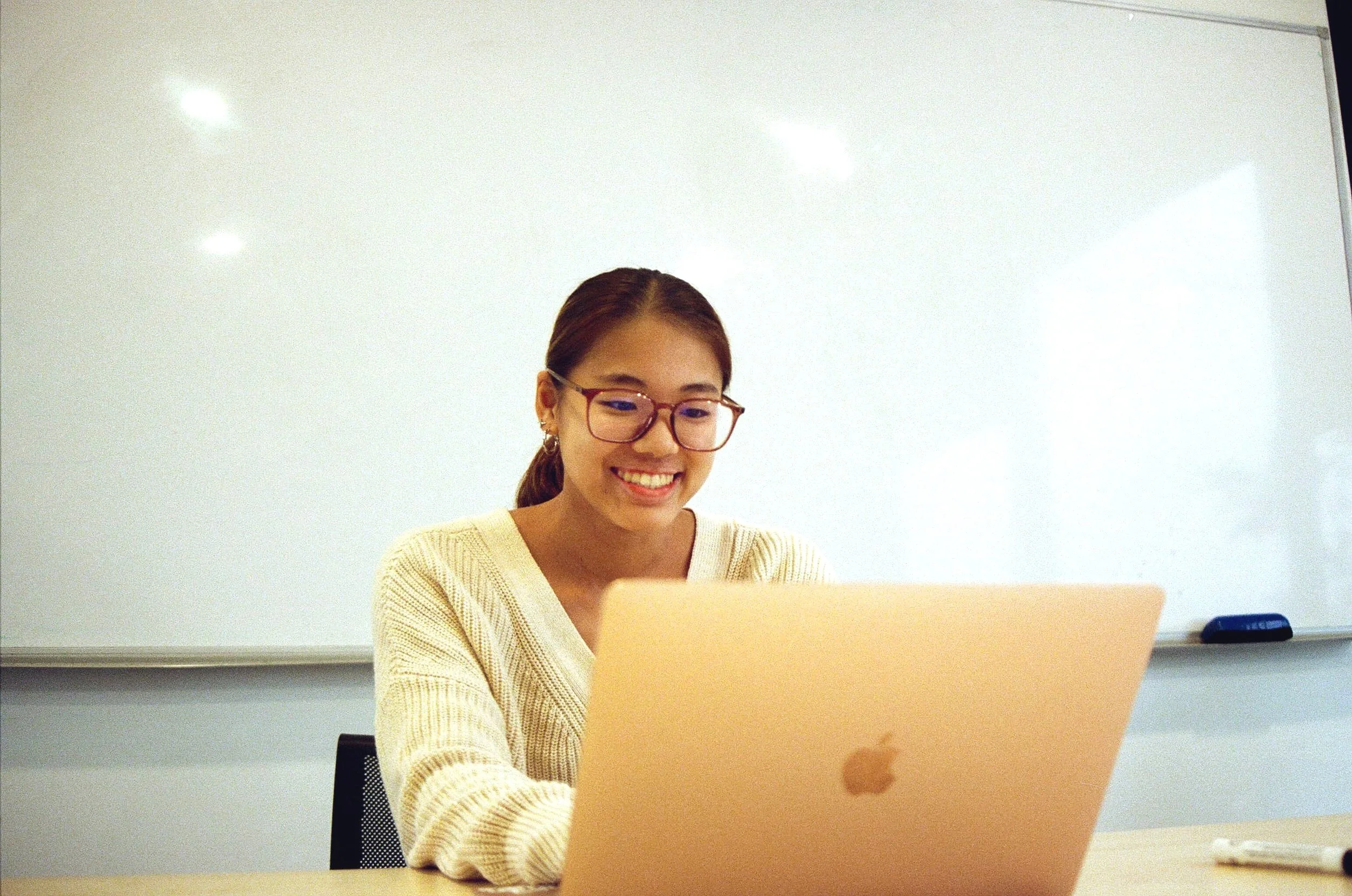 A young woman with glasses smiling while working on a laptop in a classroom or conference room with a whiteboard behind her.