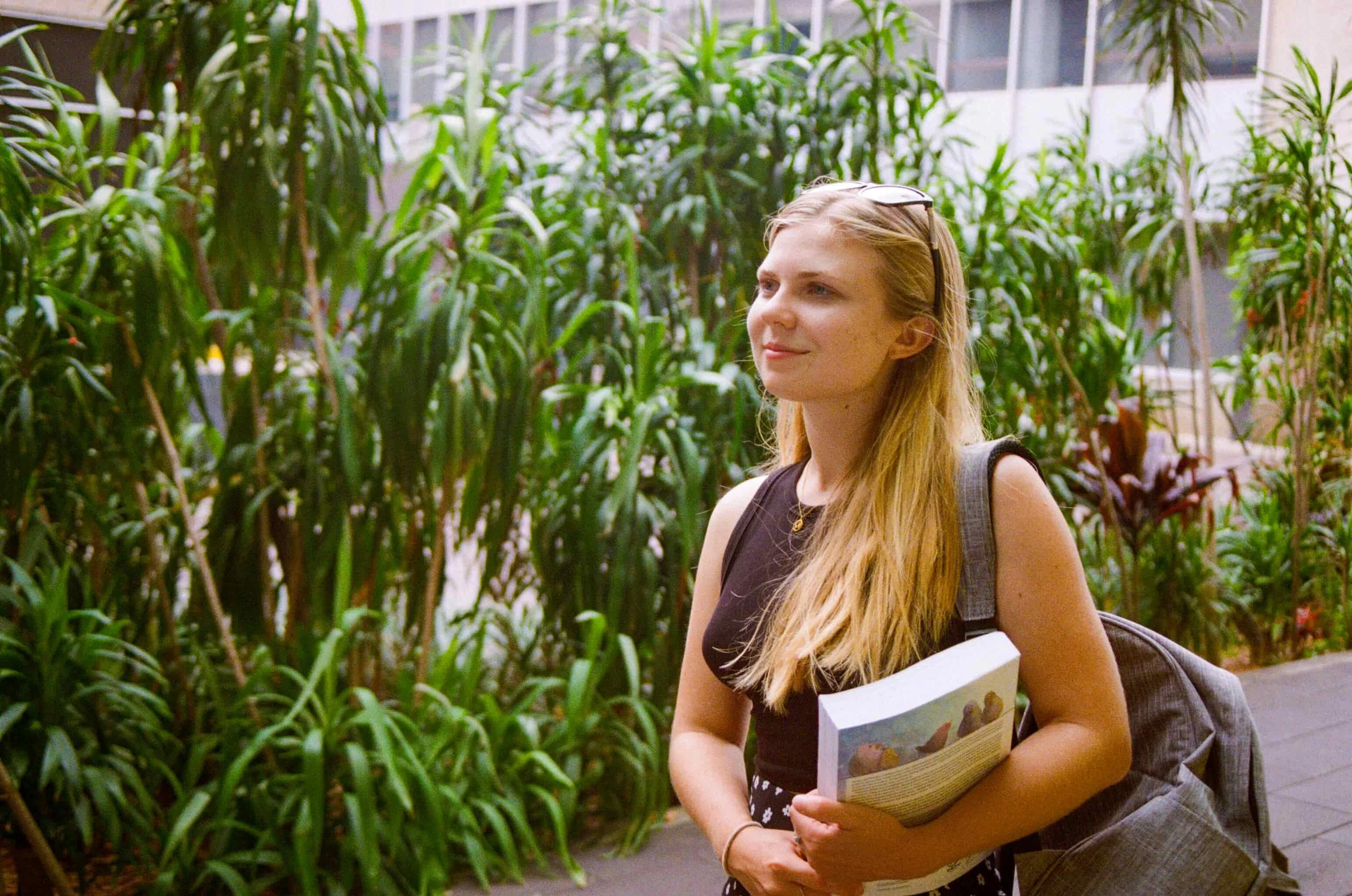 A young woman with long blonde hair, wearing a black sleeveless top, standing outdoors in front of lush green plants, holding a book and carrying a gray backpack.
