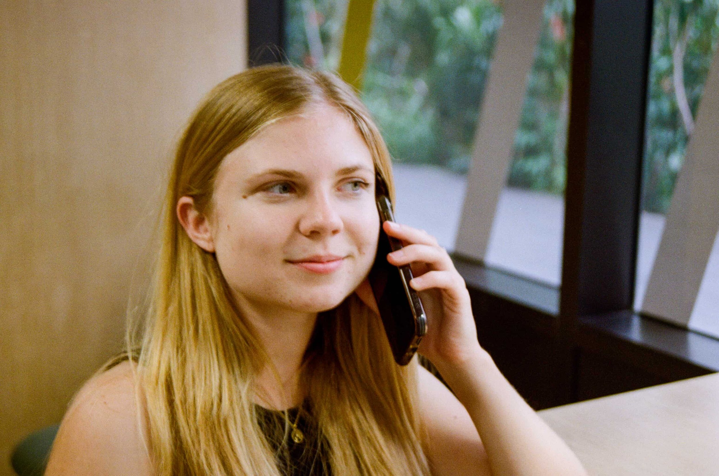 A young woman with long blonde hair is sitting indoors near a window, holding a mobile phone to her ear and smiling.