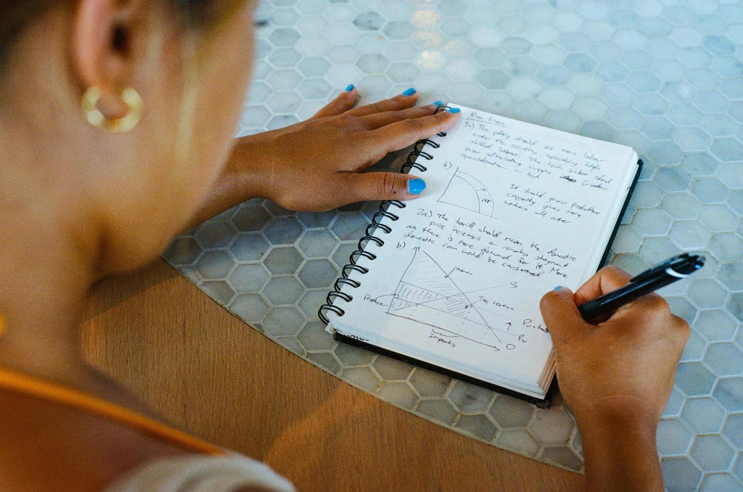 A person with blonde hair, gold hoop earrings, and blue painted nails is writing in a spiral notebook with a black pen, sitting at a table with a hexagonal tile surface and a wooden edge.