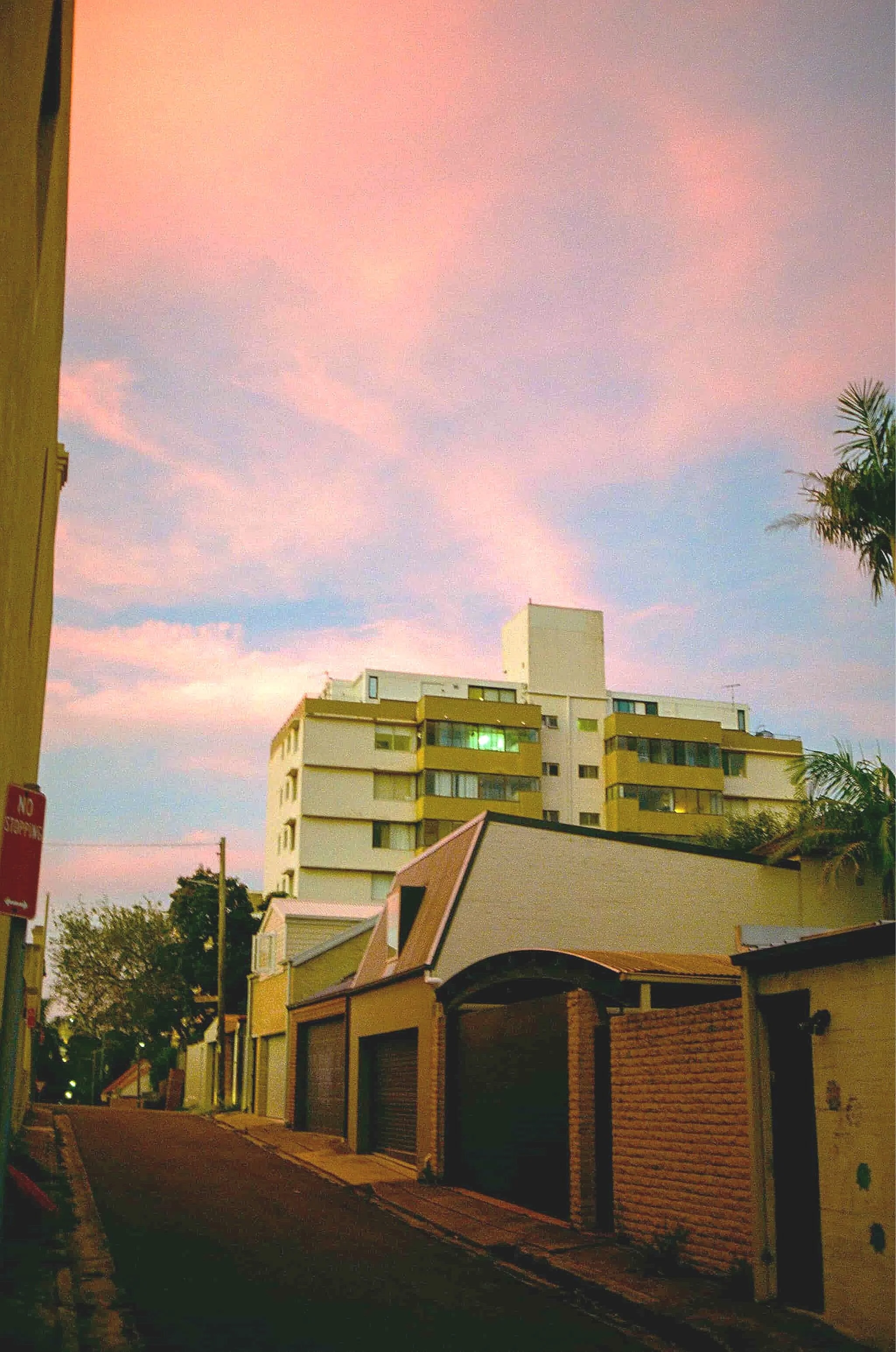 A city street scene during sunset with pastel pink and blue sky, multi-story residential buildings, and palm trees.