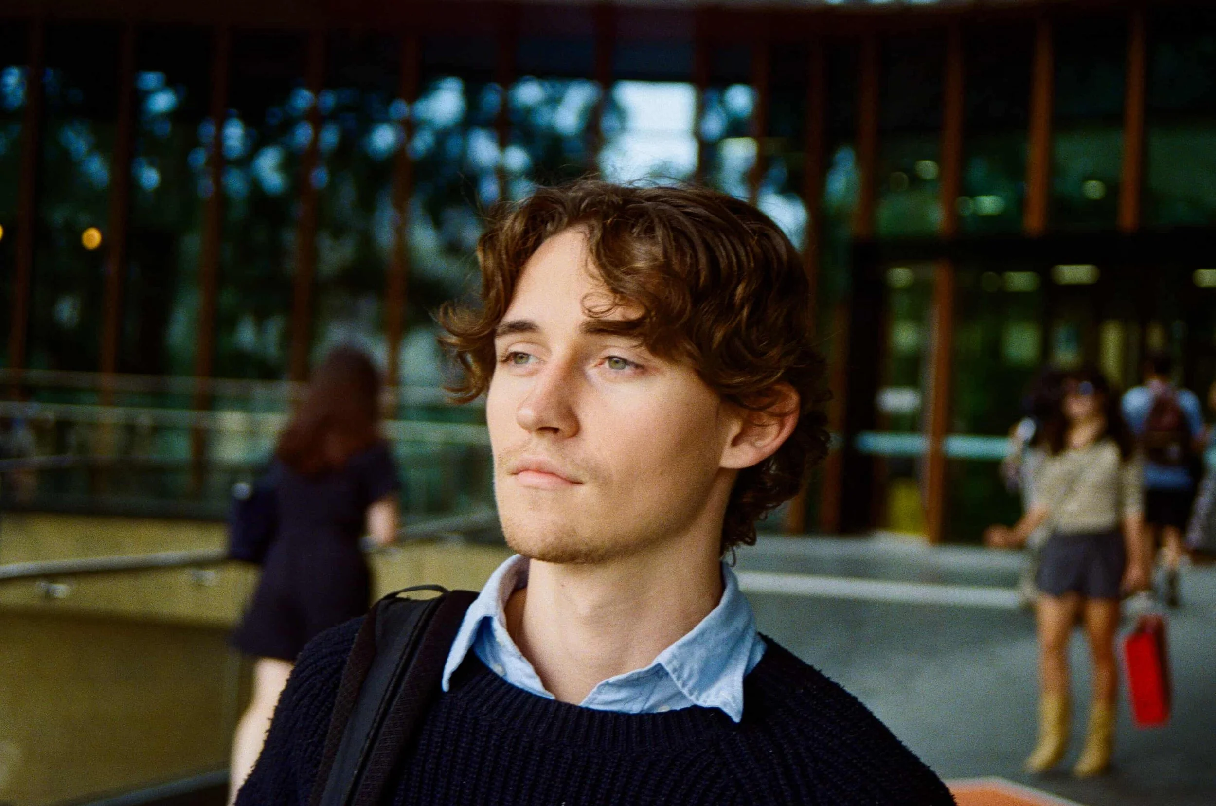 A young man with wavy brown hair, blue eyes, and light facial hair, wearing a light blue shirt and dark sweater, standing outdoors in front of a modern building with a glass facade and wooden beams. In the background, there are other people, including two women, one with sunglasses and yellow boots, and the other in black, walking and standing near the building.