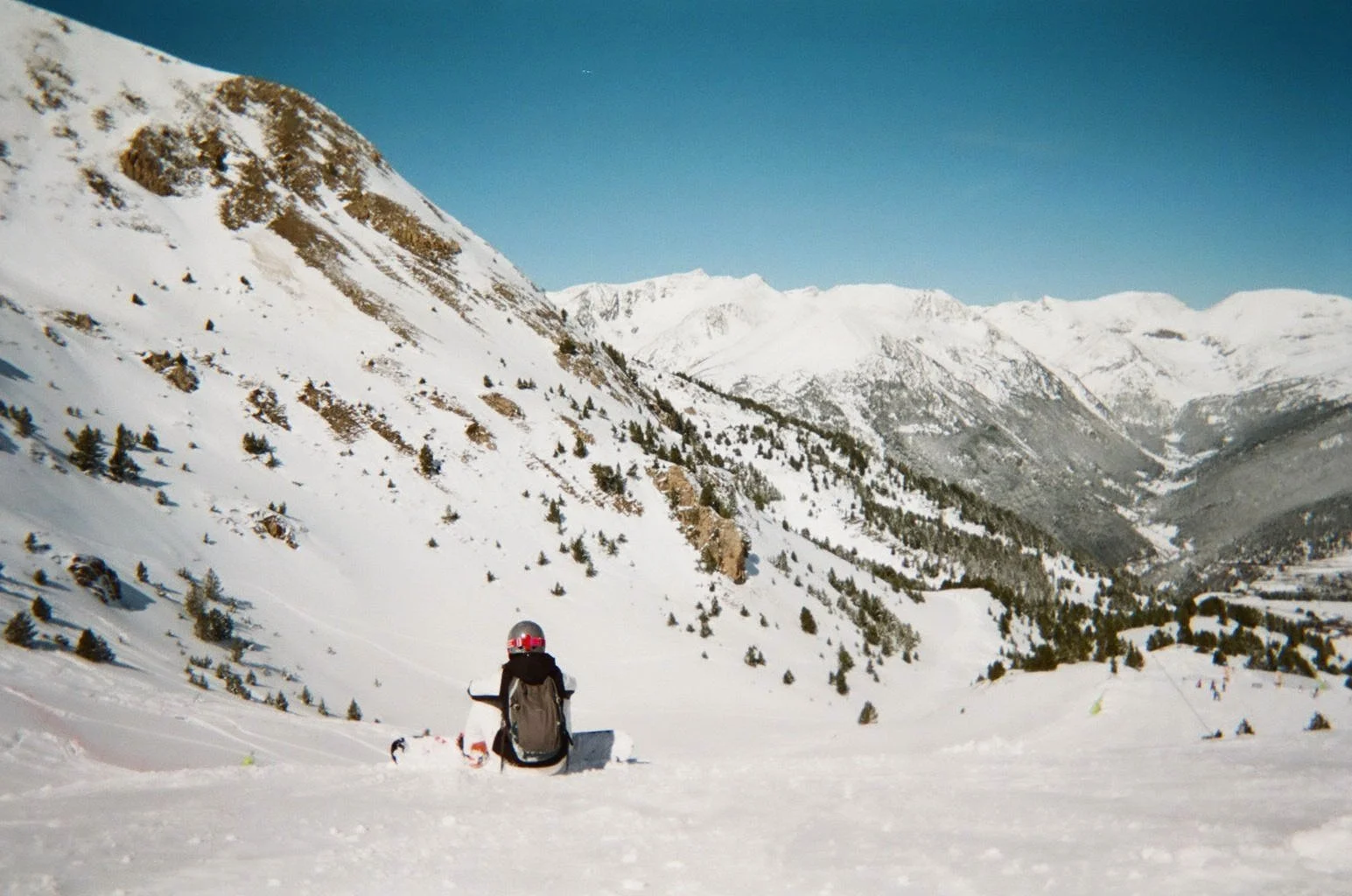A person wearing a helmet and backpack sitting on snowy ground, gazing at snow-covered mountains and a valley under a clear blue sky.