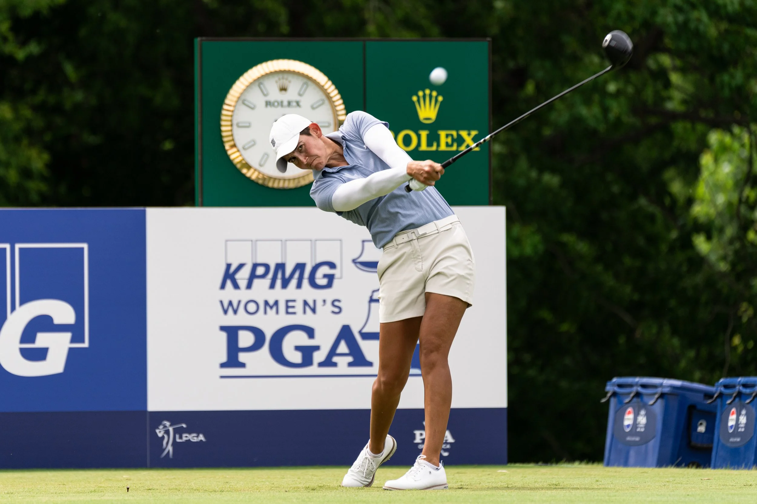 Young woman in golf attire swinging a golf club on a golf course, with a focused expression.