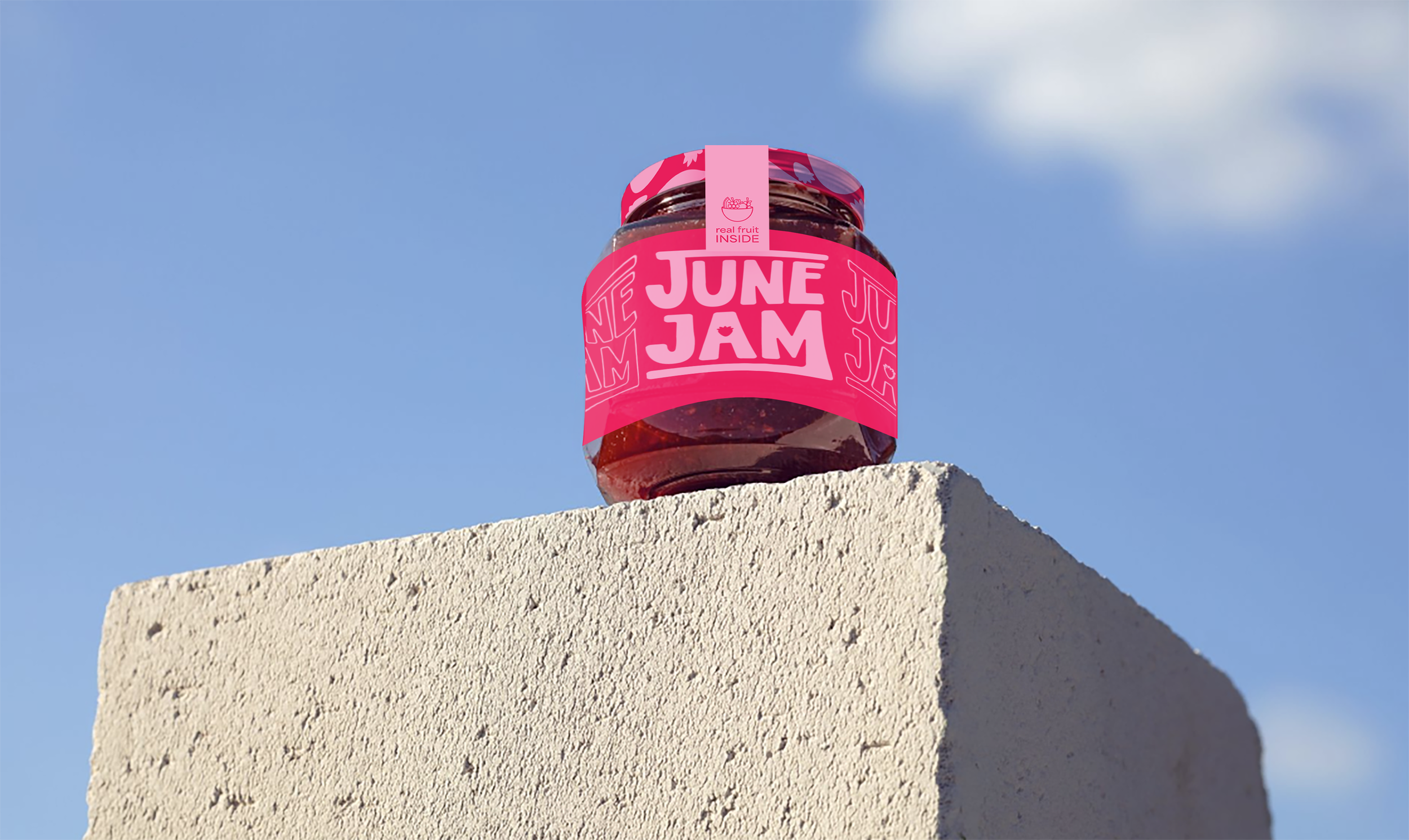 Image of a jar of June Jam's strawberry Guava flavoured jam. The jar has pink branding with strawberry illustrations. The jar is sitting on top of a grey cinderblock with a blue sky behind it.