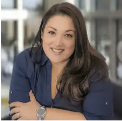 A woman smiling with long dark hair wearing a blue shirt in an indoor setting.