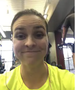 A woman with her hair pulled back, wearing a bright yellow shirt, smiling at the camera inside a gym or fitness center.