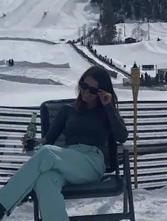A woman sitting outdoors in snow, wearing sunglasses, a dark long-sleeve shirt, and light blue pants, with a snow-covered landscape and ski lift in the background.