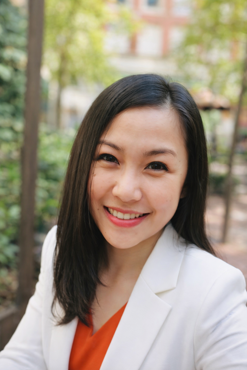 Smiling woman with dark hair wearing a white blazer and orange top, outdoors with greenery in the background.