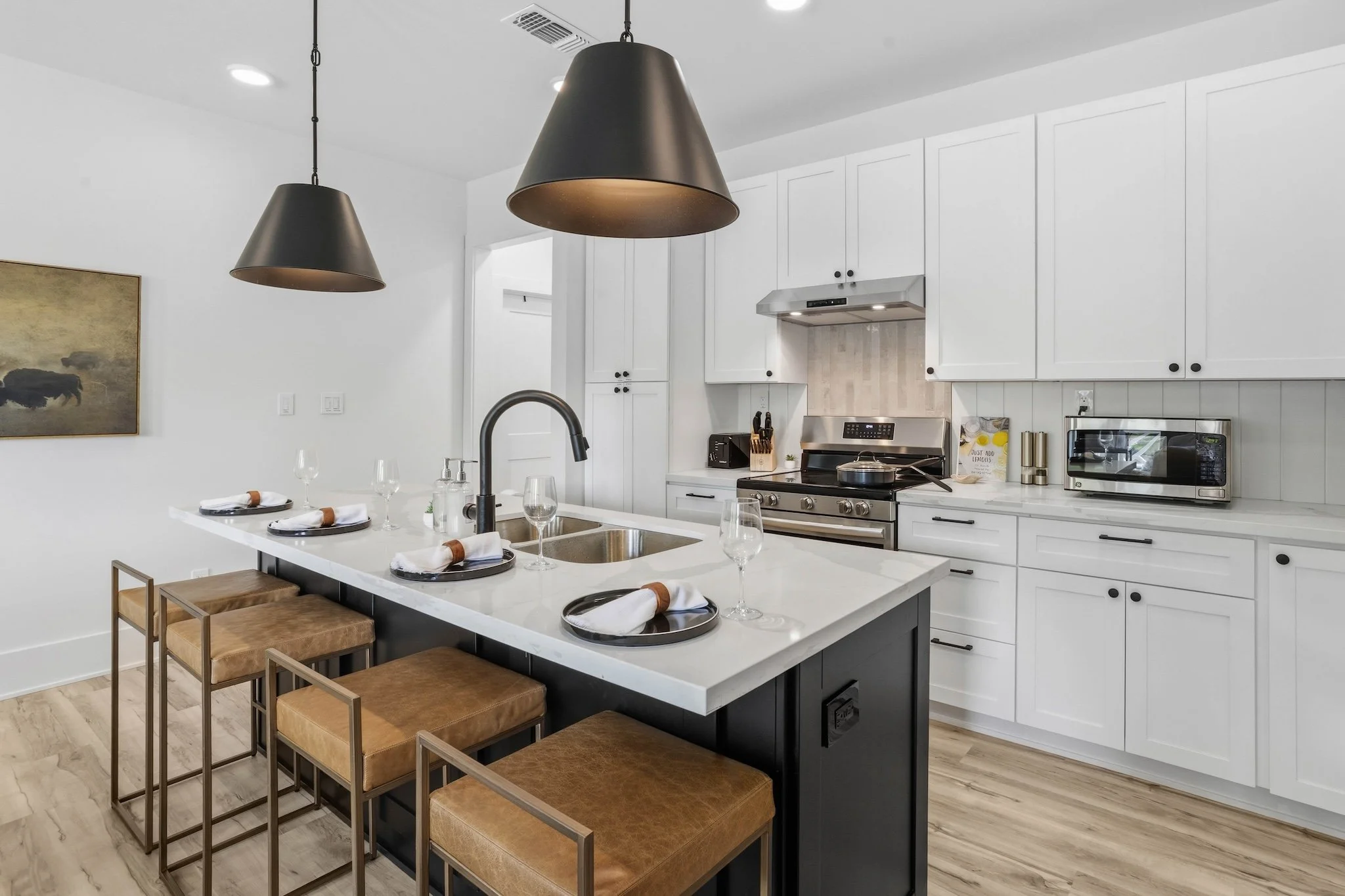 Modern kitchen with white cabinets, a black island, and black pendant lights. The island has four place settings with napkins, plates, and glasses. The kitchen includes a stainless steel stove, microwave, and utensils. Light wood flooring and a wall 
