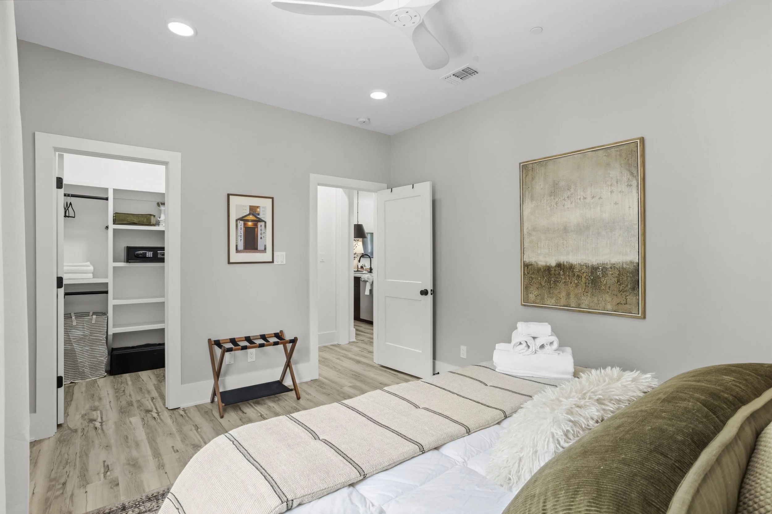 A bedroom with a bed covered in a beige striped blanket and decorative pillows. A stack of white towels is on the bed. There's a large abstract painting on the wall, and a wooden luggage rack near the wall. The room has wood flooring, a closet with s
