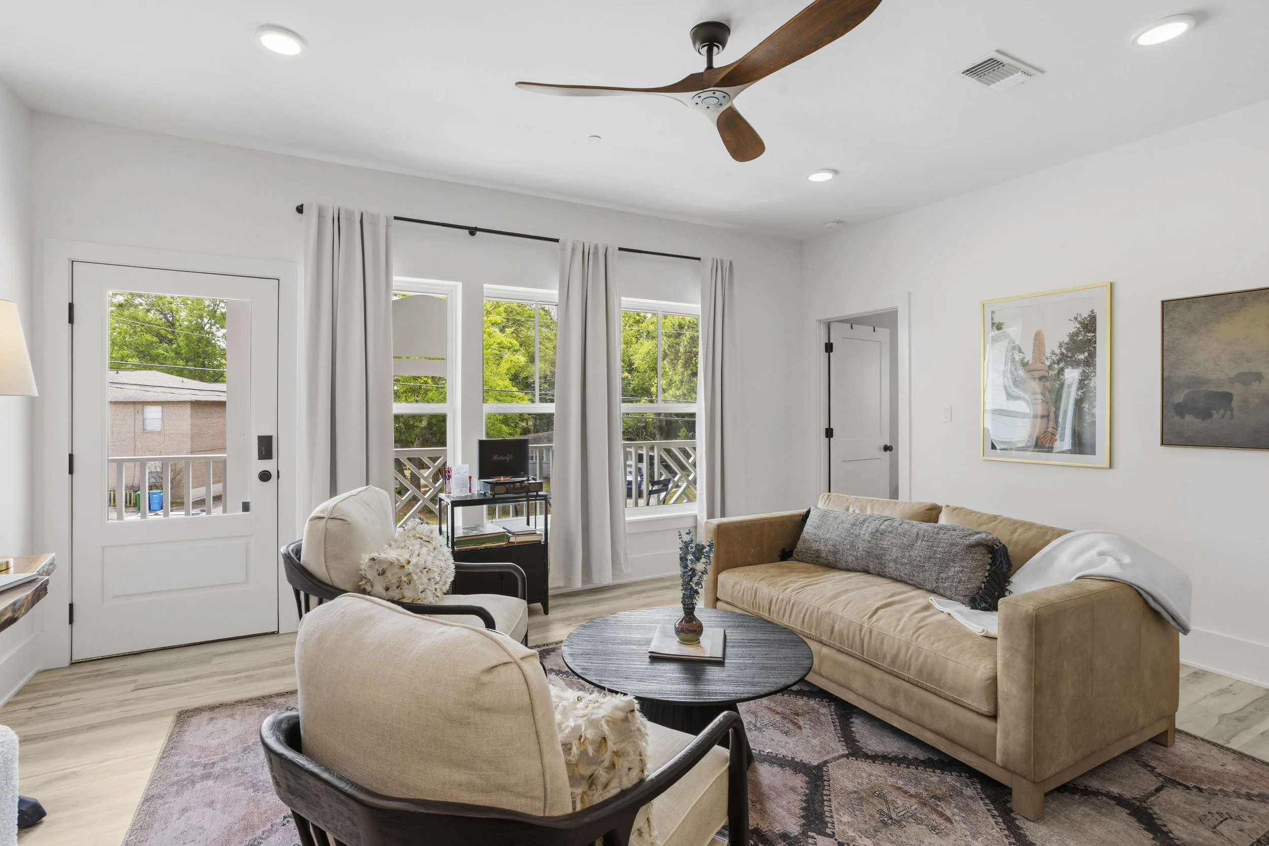 Living room with beige sofa, two beige armchairs, black round coffee table, white walls, three windows with white curtains, wooden ceiling fan, artwork on wall, hardwood floor, porch visible outside.
