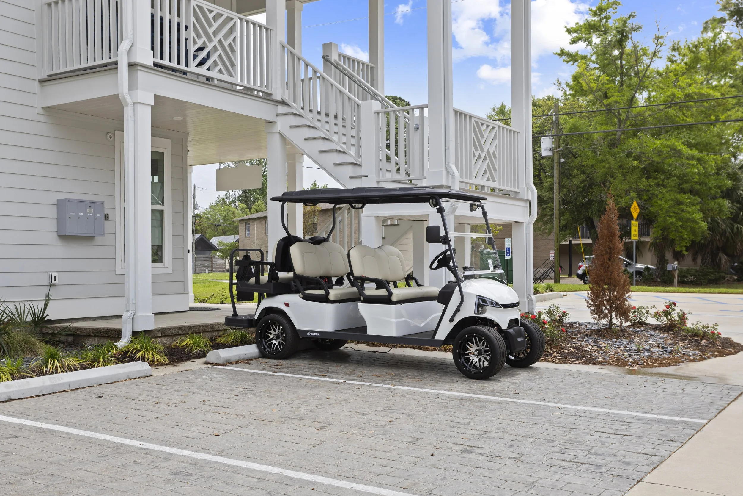 A white electric golf cart parked in a residential parking lot in front of a light gray house with white trim and a wooden staircase leading to a second-story balcony.