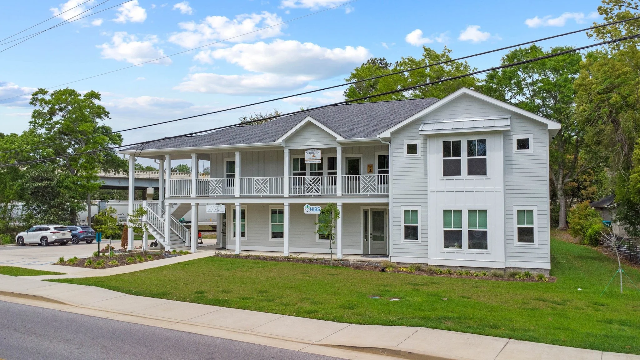 A modern two-story building with a large balcony and white siding, surrounded by green grass and trees, with parked cars nearby.