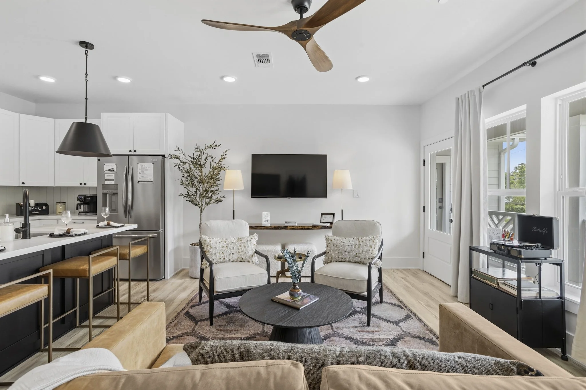 Living room with white walls, two chairs with pillows, a black coffee table with a decor item, a flat-screen TV on the wall, two floor lamps, curtains, a sliding glass door, and a kitchen area with barstools.