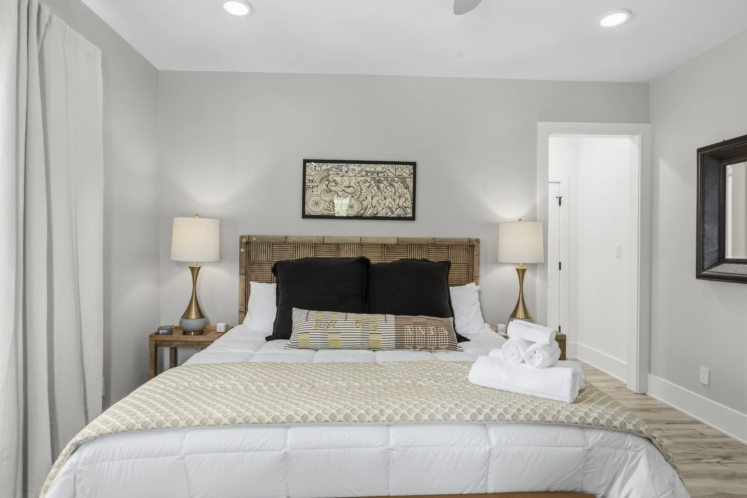 A neatly made bed with beige and white bedding, four rolled white towels on the bed, and black and beige decorative pillows. Two matching gold bedside lamps are on wooden nightstands on either side of the bed. The bedroom has a gray wall, a framed ar