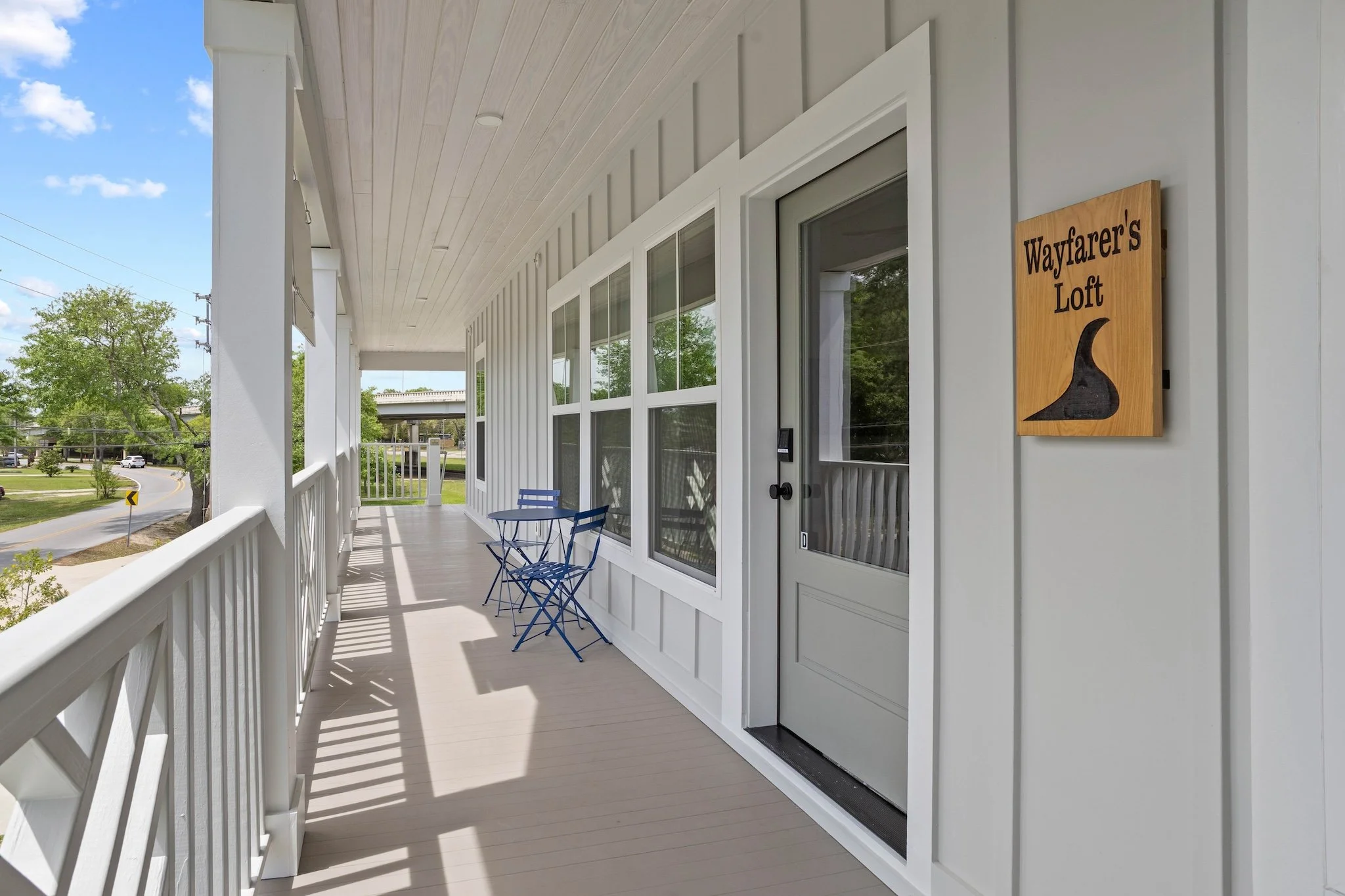 A porch with a white railing and wall, two blue folding chairs, and a sign that reads "Wayfarer's Loft" with a black hook-shaped graphic.