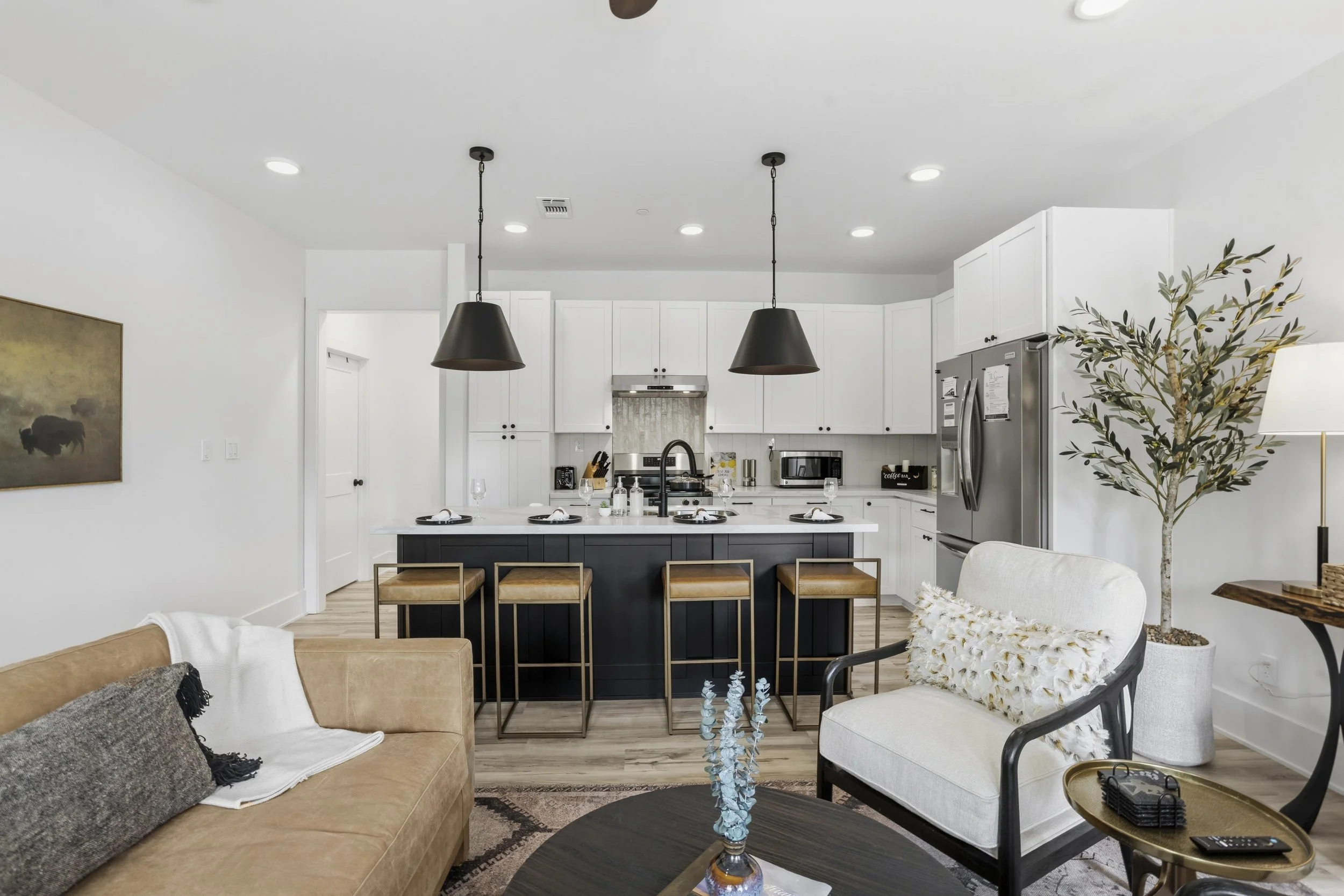 Open concept living room and kitchen with white cabinetry, black island, three black pendant lights, beige sofa, white and black armchair, large potted plant, and decorative items.