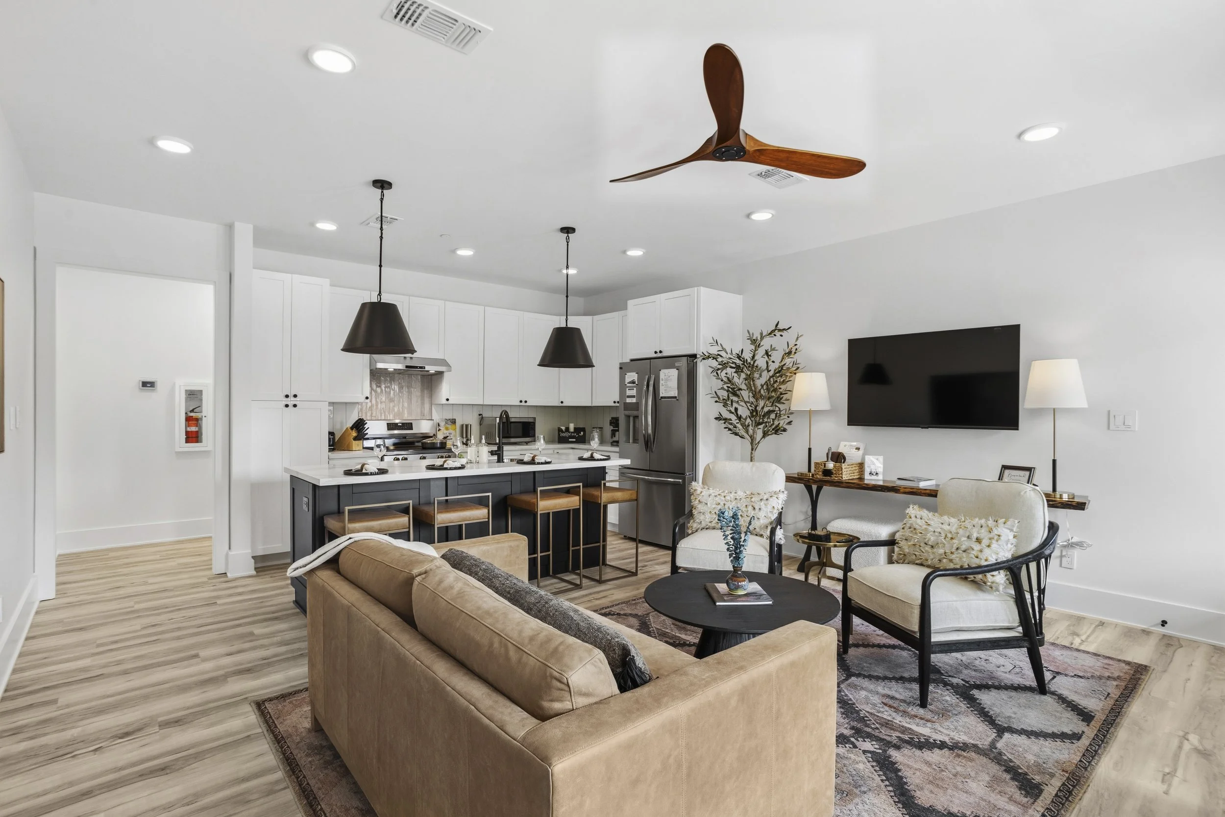 Open-concept living room and kitchen with a beige sofa, white and black armchairs, a black round coffee table, and a wall-mounted TV.