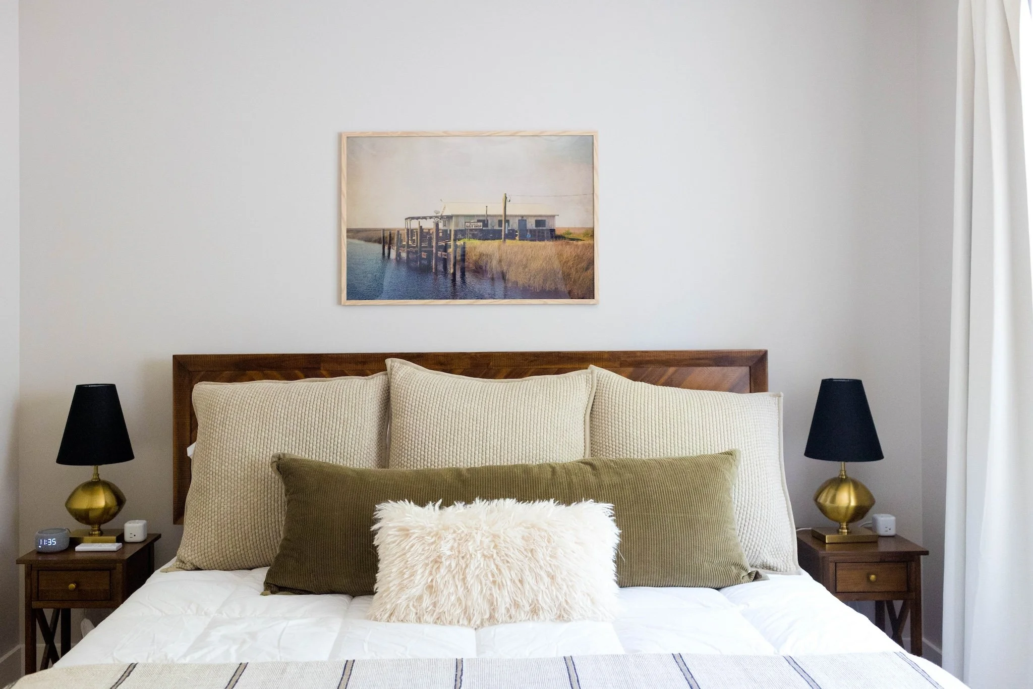 A bedroom with a wooden headboard, cream and green pillows, and a white fluffy pillow. Two black lamps with gold bases on wooden nightstands on each side. A framed photo of a dock and water hangs above the bed. White curtains are partially visible on the right.