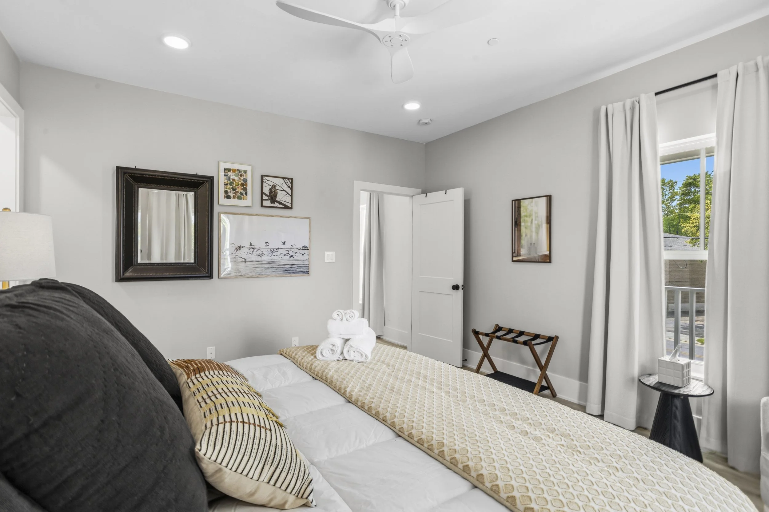 A neatly made bed with white linens, a beige patterned blanket, and striped pillows in a bright bedroom with modern decor, framed wall art, a window with white curtains, and a small side table.