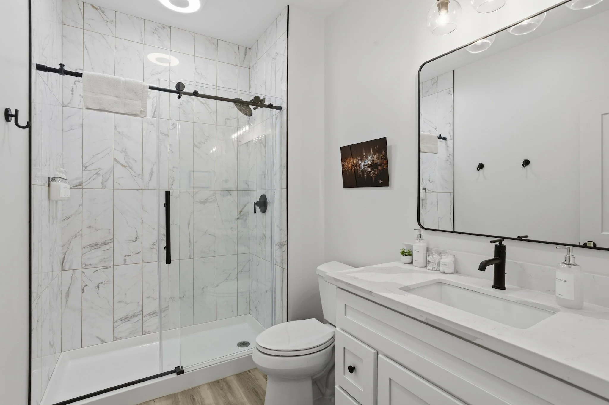 Modern bathroom with a glass-enclosed shower with marble tiles, white vanity with a marble countertop, large mirror, black fixtures, and minimal decor.