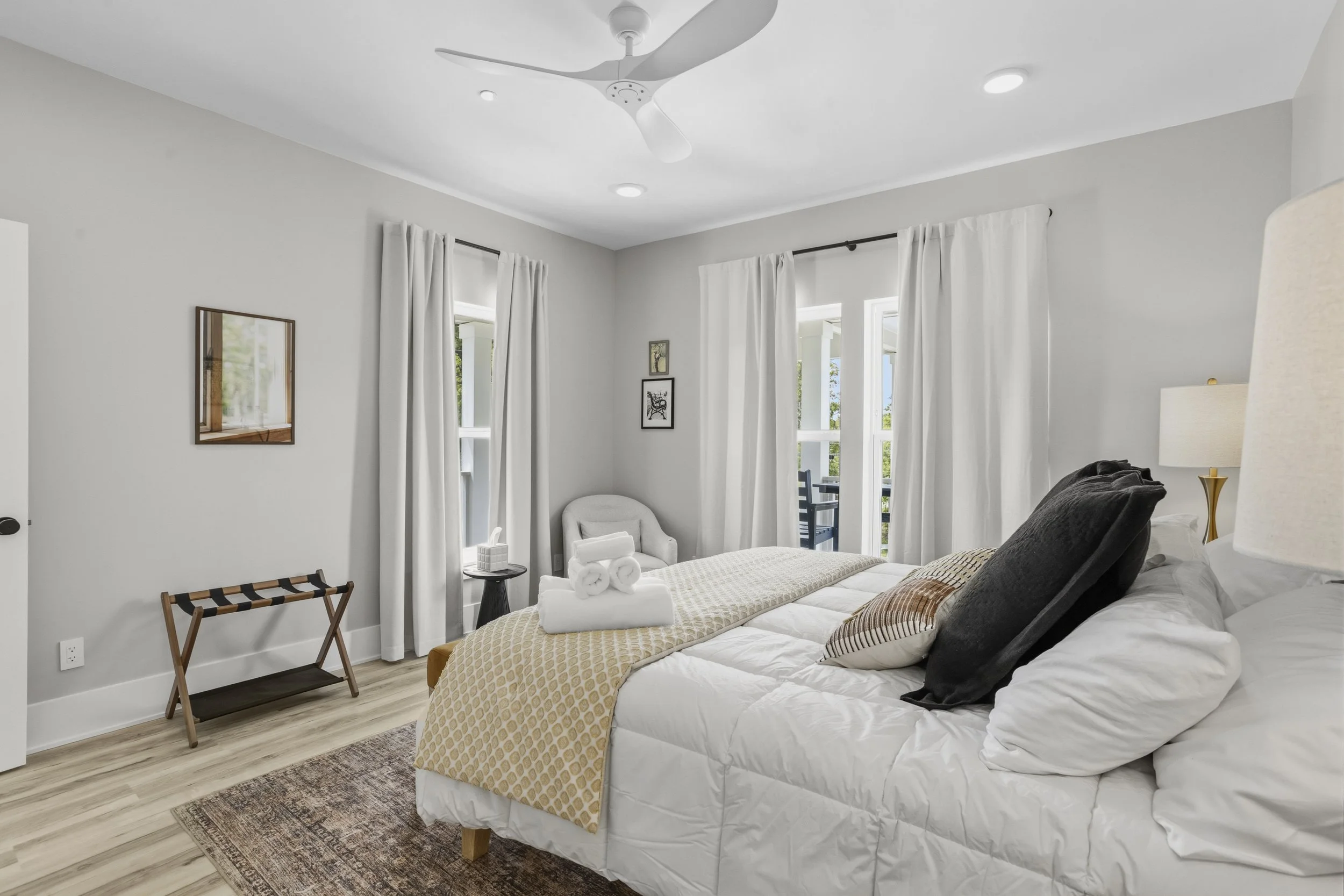 A bedroom with white walls, a bed with white linens and black pillows, a bedside lamp, a small sitting chair, a window with white curtains, a ceiling fan, and wall art.