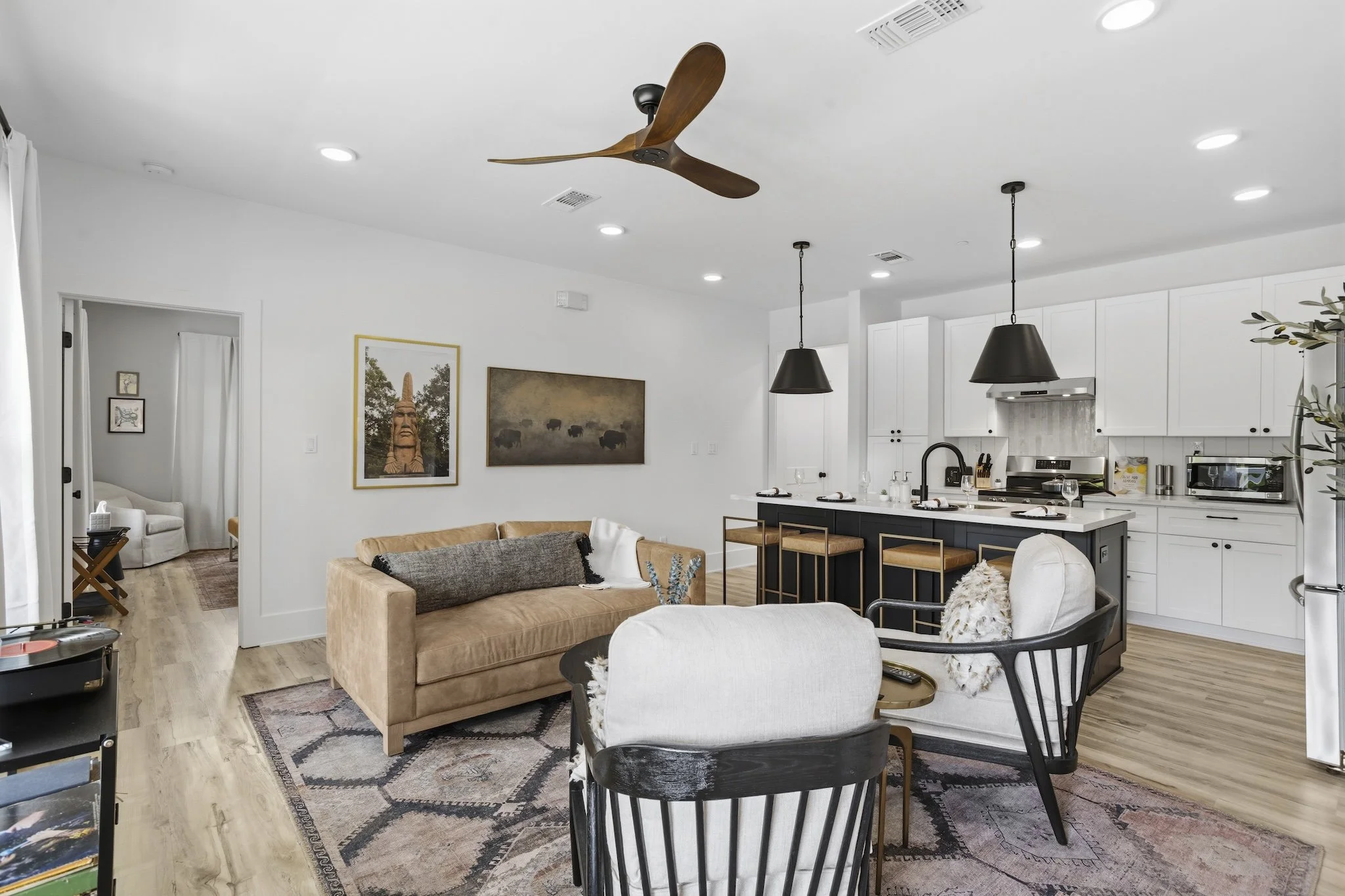Open-concept living room and kitchen with white cabinets, black accents, and wooden furniture, ceiling fan, and artwork.