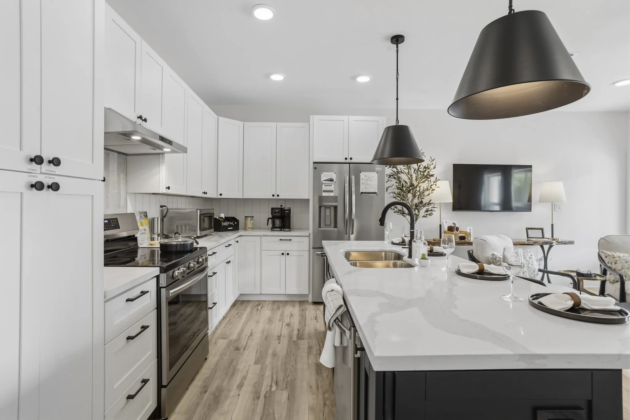 Modern kitchen with white cabinets, stainless steel appliances, a large island with a white quartz countertop, black pendant lights, and a view of the living room area with a wall-mounted TV, plants, and seating.