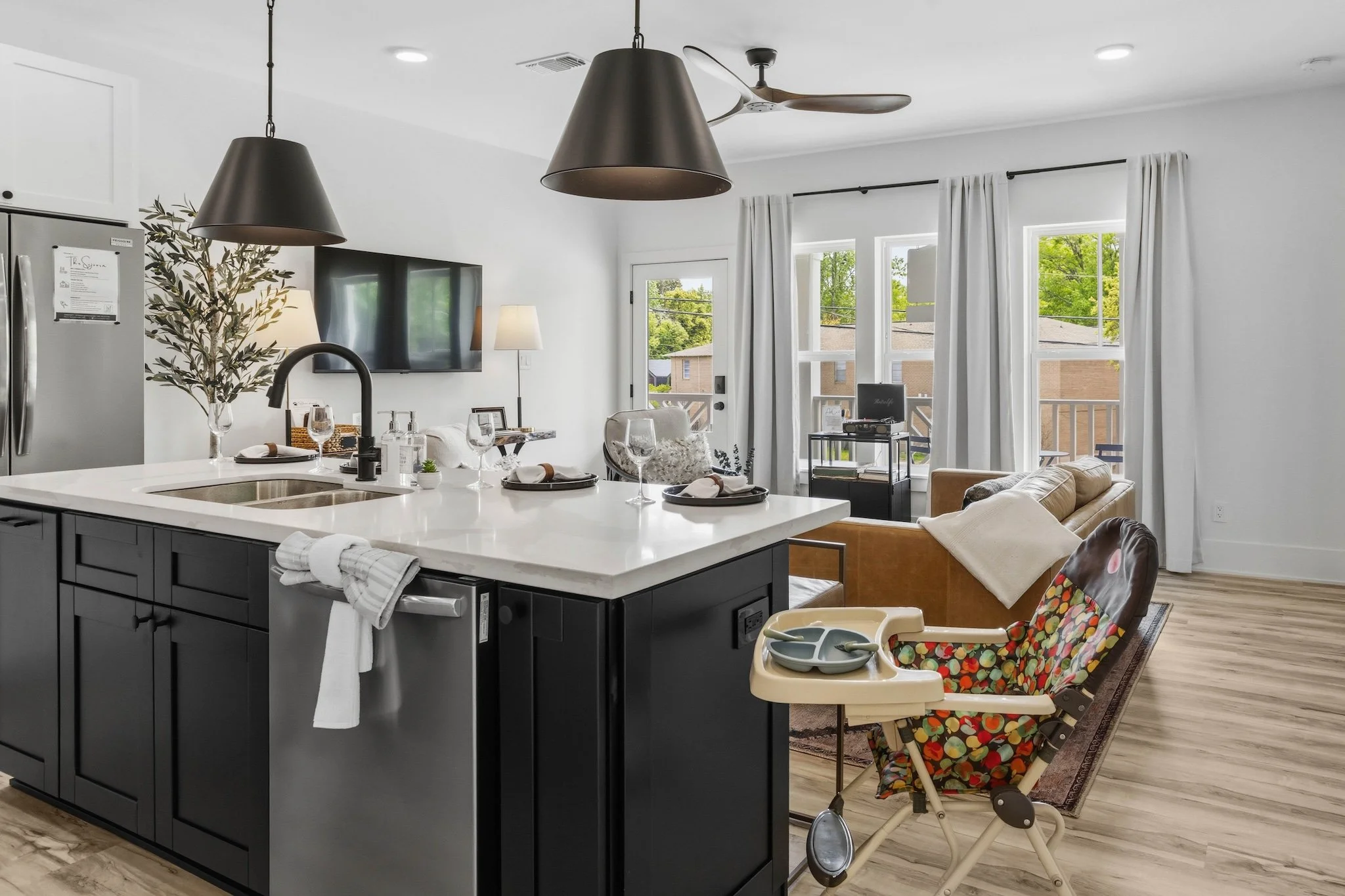 Open-concept living room and kitchen with white walls, large windows with white curtains, ceiling fan, and two black pendant lamps over a white kitchen island. The island has a sink, wine glasses, and table settings. A high chair with a colorful polk