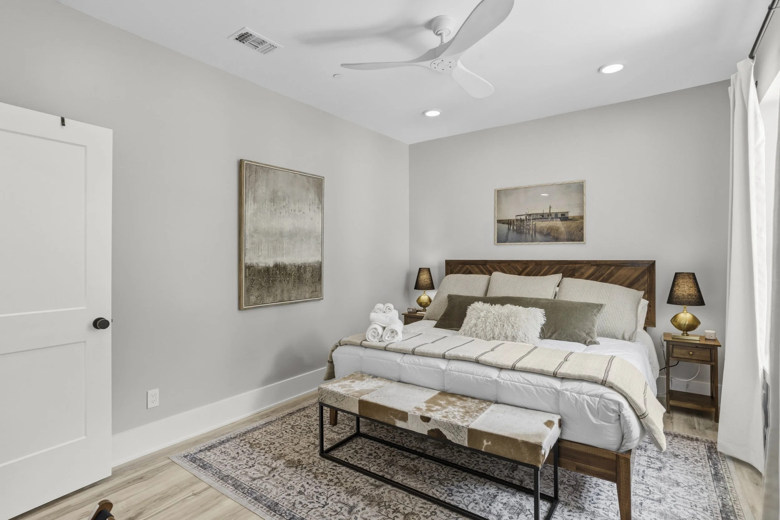 Modern bedroom with wooden bed frame, beige pillows, a bench with cowhide, framed artwork, and nightstands with black lamps, white curtains, and wood flooring.