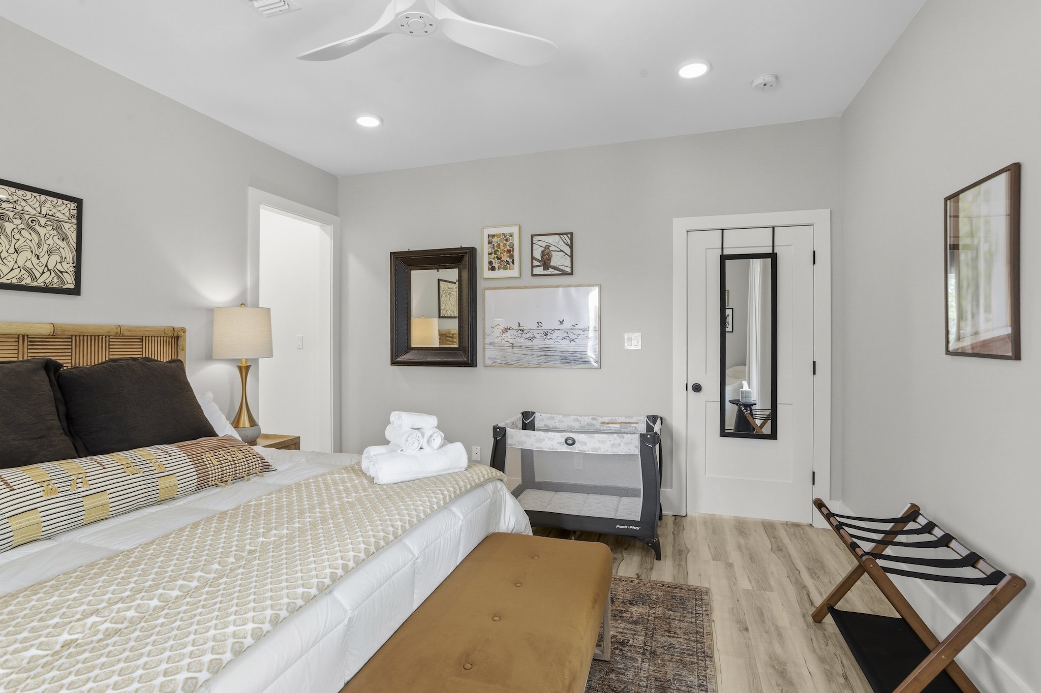 A bedroom with a bed, nightstand, lamp, art on the wall, a mirror, a pack and play, a folding luggage rack, and a wall-mounted mirror.
