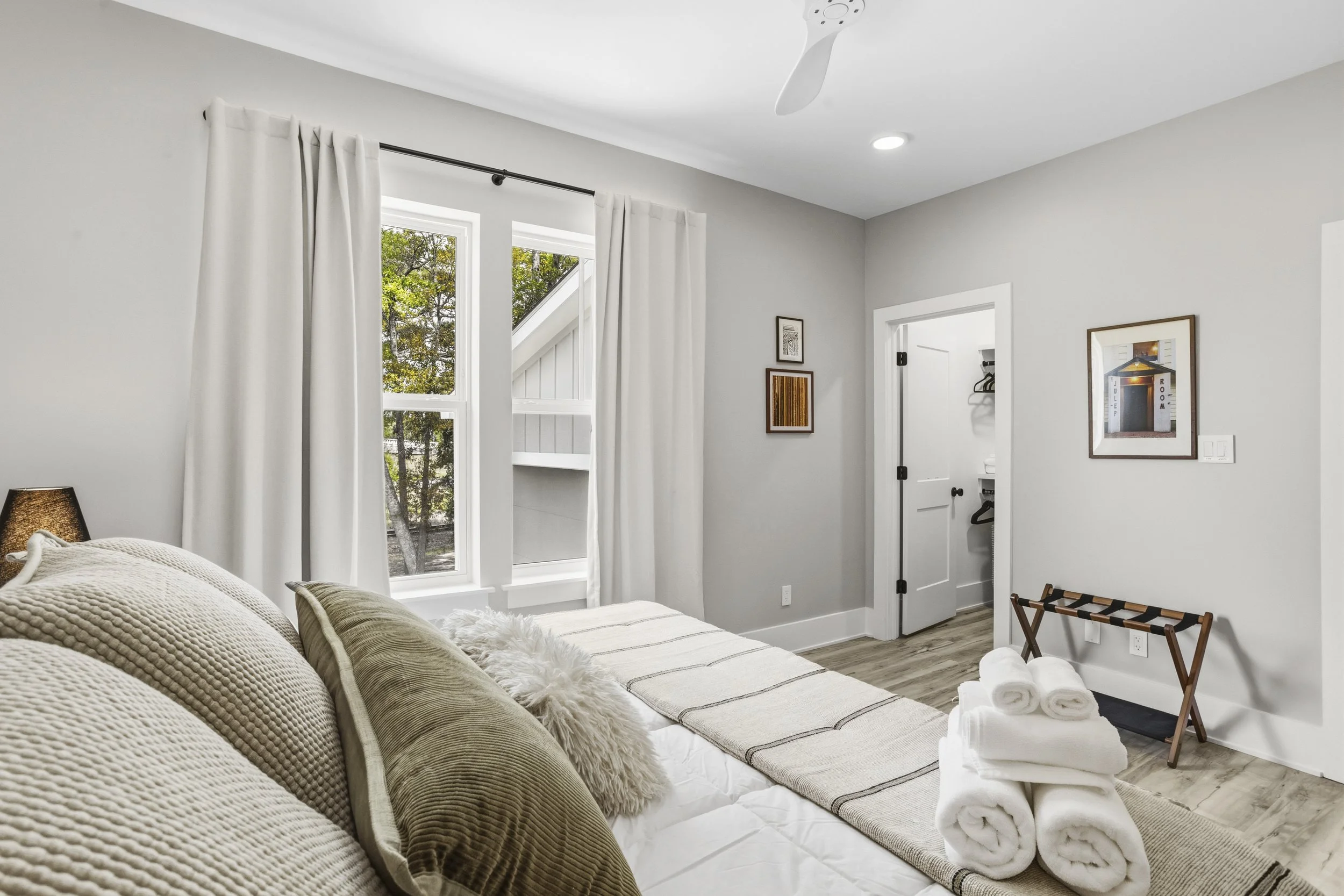 Bright bedroom with large windows, white curtains, neutral-colored bedding, and decorative pillows. Small art pieces hang on light gray walls. Towels are neatly rolled on a small bench. A door leads to a closet, and a ceiling fan and recessed lightin
