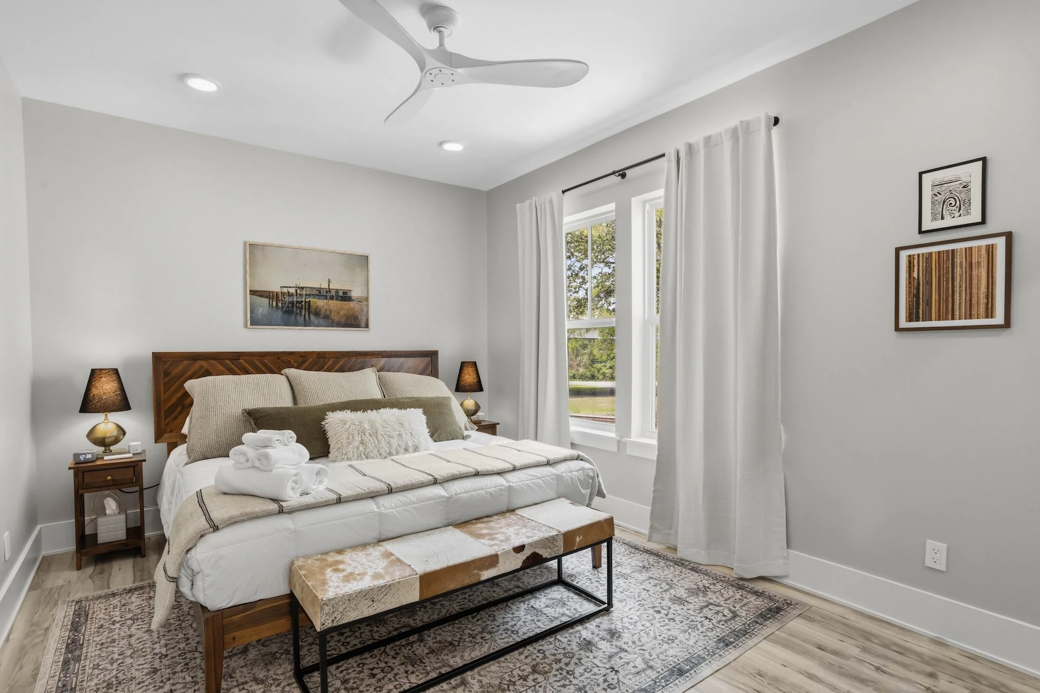 A bedroom with a wooden bed, white bedding, and multiple pillows, including a fluffy white pillow and a green rectangular pillow. There are two nightstands with black lamps on each side of the bed, a framed artwork above the headboard, a bench at the