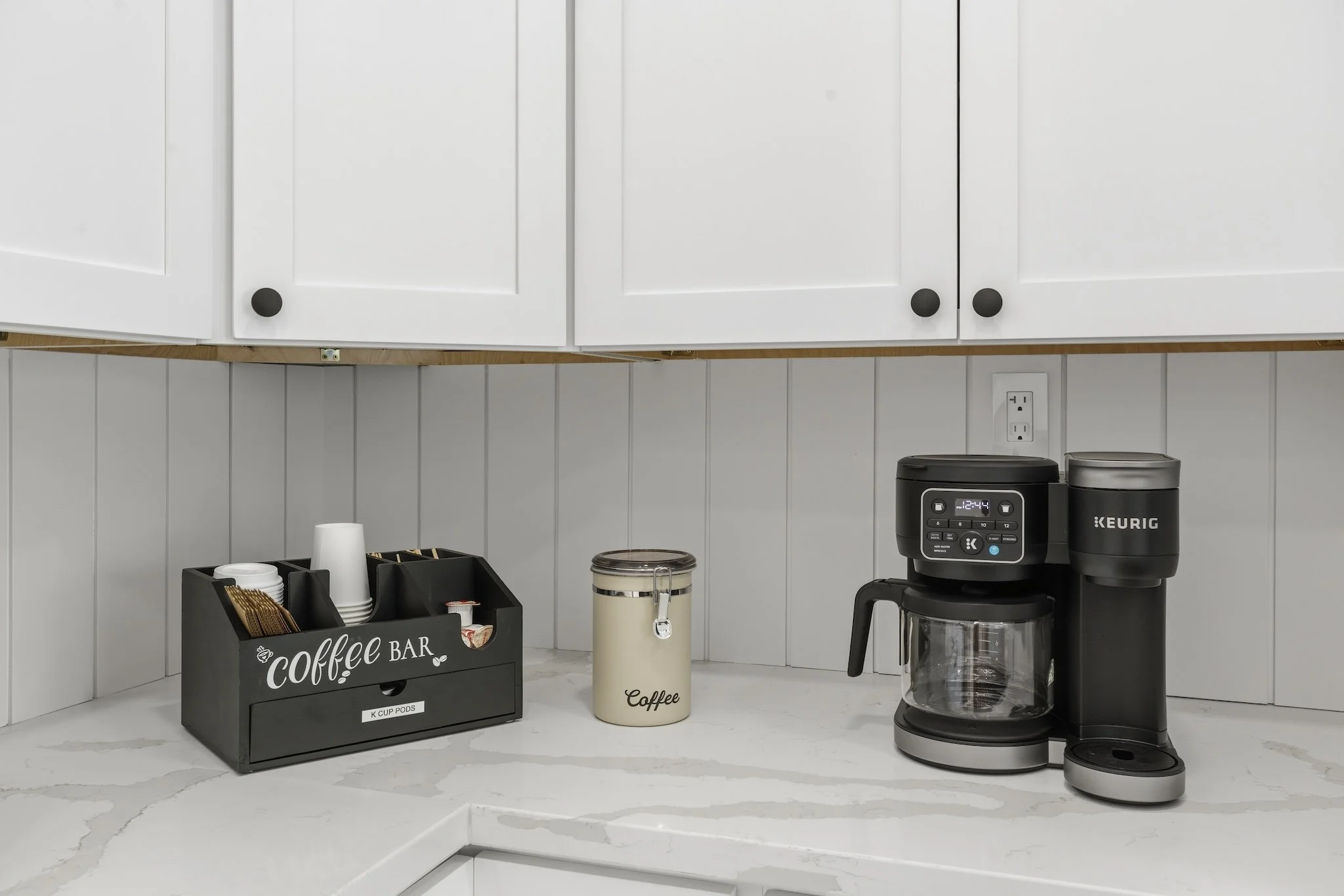 Coffee station with black coffee maker, white cabinets, and counter with coffee supplies.