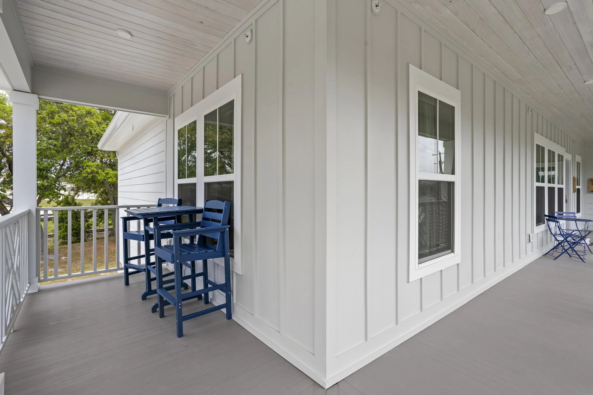 A spacious house porch with white siding, four windows, and light gray flooring. Blue outdoor furniture and a small table set are present, with a view of green trees and a neighbor's house in the background.