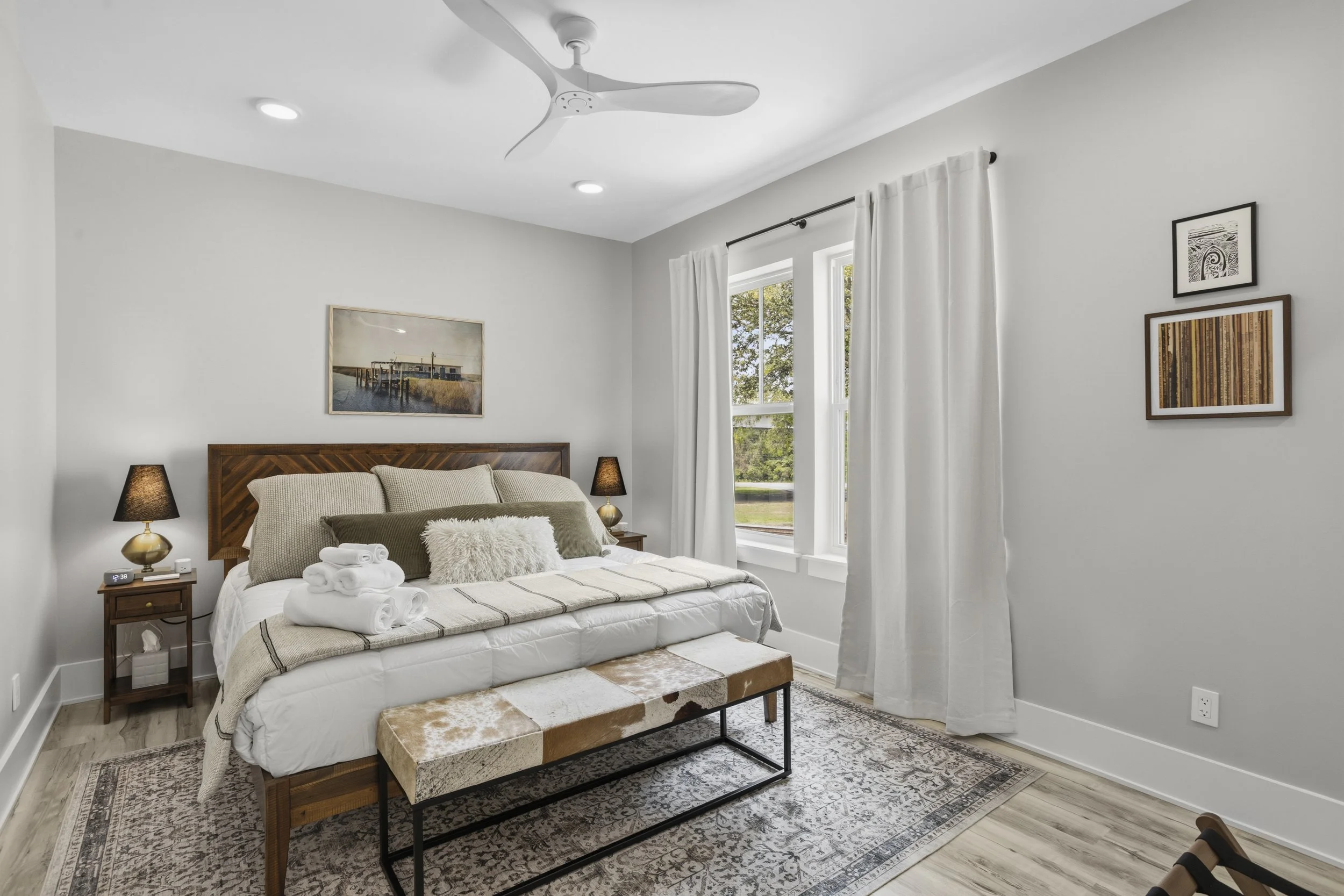 A neatly made bedroom with a white bed, beige pillows, a furry accent pillow, and white rolled towels on top. There are two small wooden nightstands with black lamps flanking the bed. The room has light gray walls, white curtains over a window showin