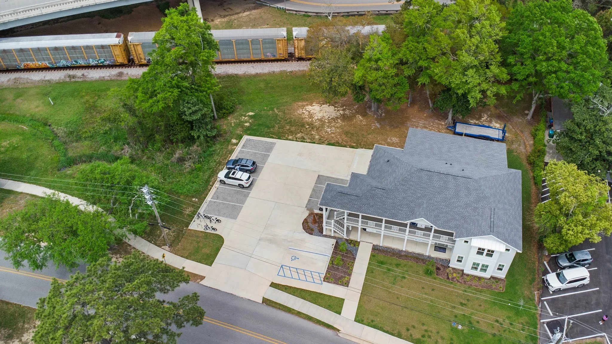Aerial view of a house with a large driveway including parking spaces, bicycles, surrounded by trees and green grass, with a railway and train cars nearby.