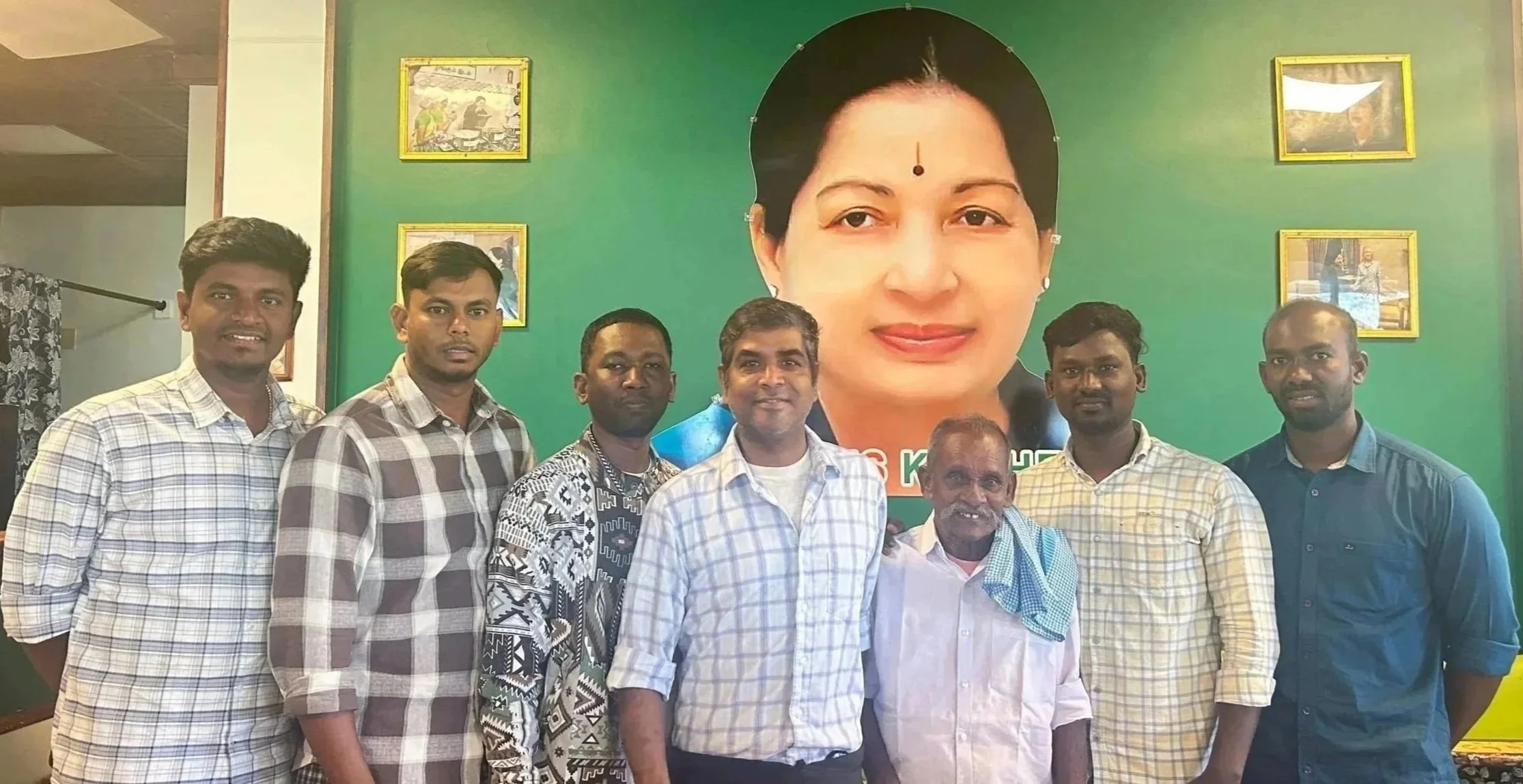 A group of eight men standing in front of a large portrait of a woman on the wall, in an indoor setting.