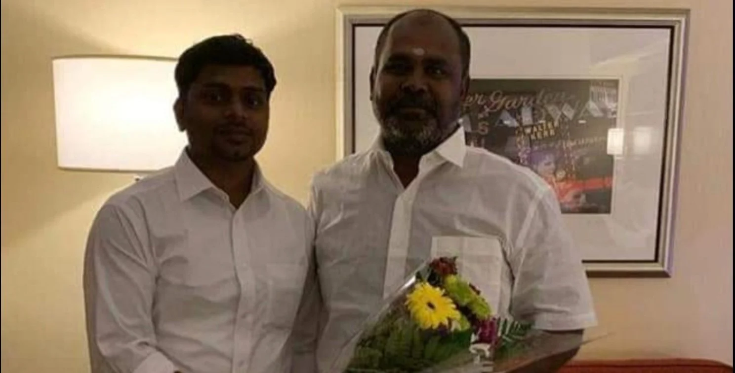 Two men in white shirts standing indoors, one holding a bouquet of flowers, with a framed picture on the wall behind them.