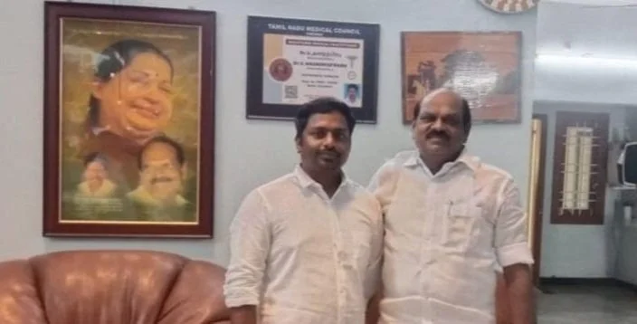 Two men standing side by side in an office, smiling. Behind them are framed pictures and documents on the wall, including a portrait of a woman and a certificate.