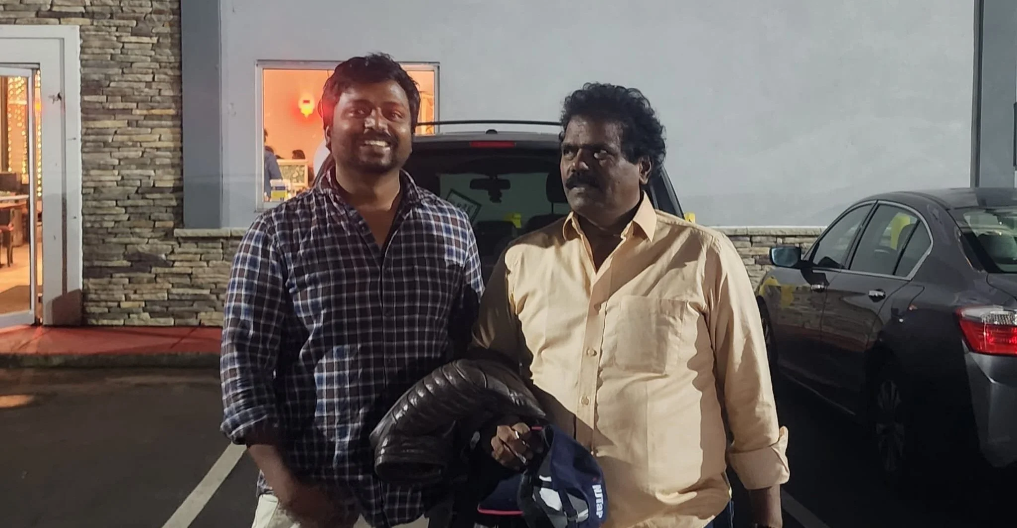 Two men standing outside a restaurant at night, with parked cars behind them. One man is smiling, wearing a checkered shirt, and the other has a serious expression, wearing a light-colored shirt and holding a motorcycle helmet.