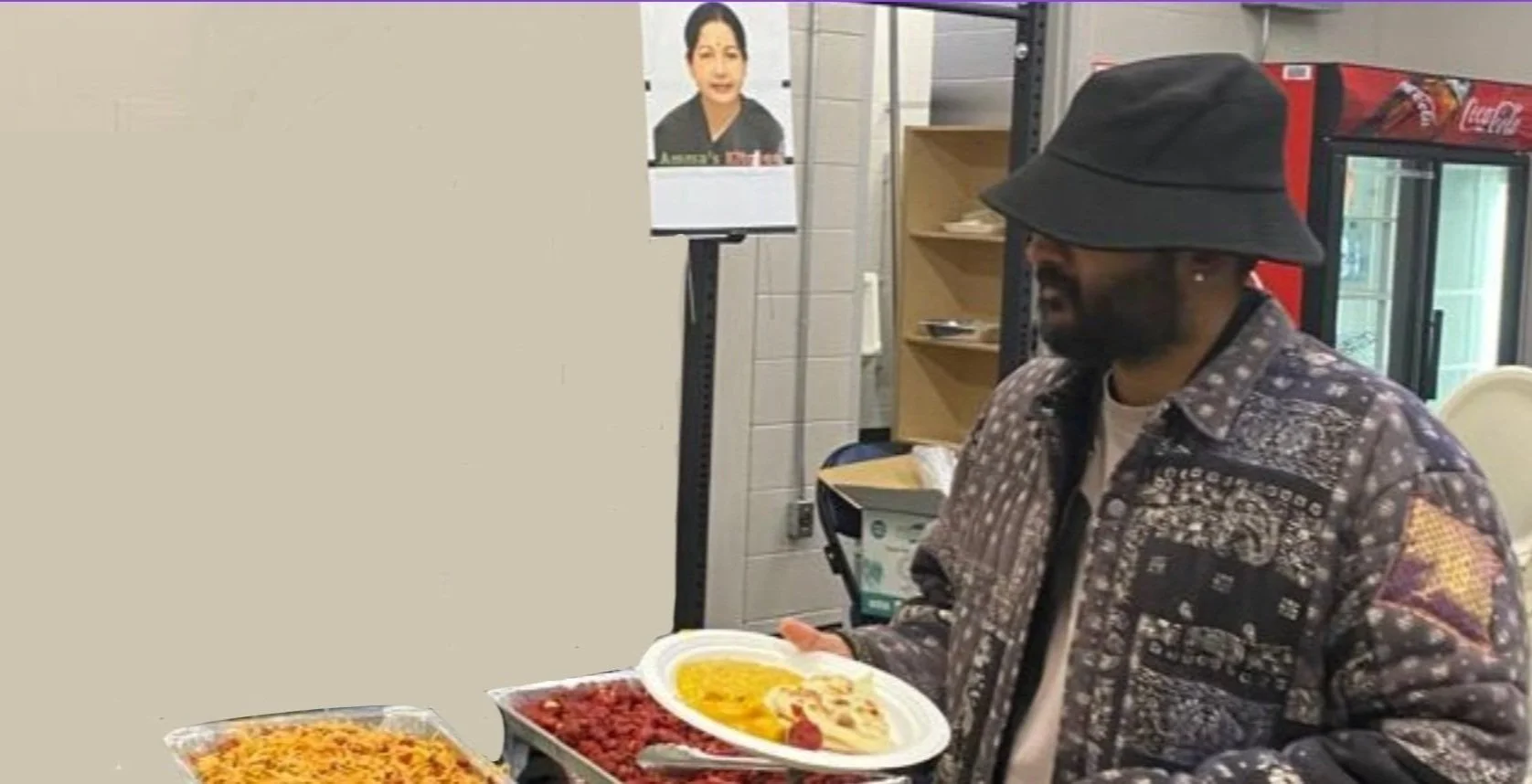 A man wearing a black hat and patterned jacket holding a plate of food in a cafeteria or restaurant setting with a poster of a woman on the wall and a Coca-Cola refrigerator in the background.