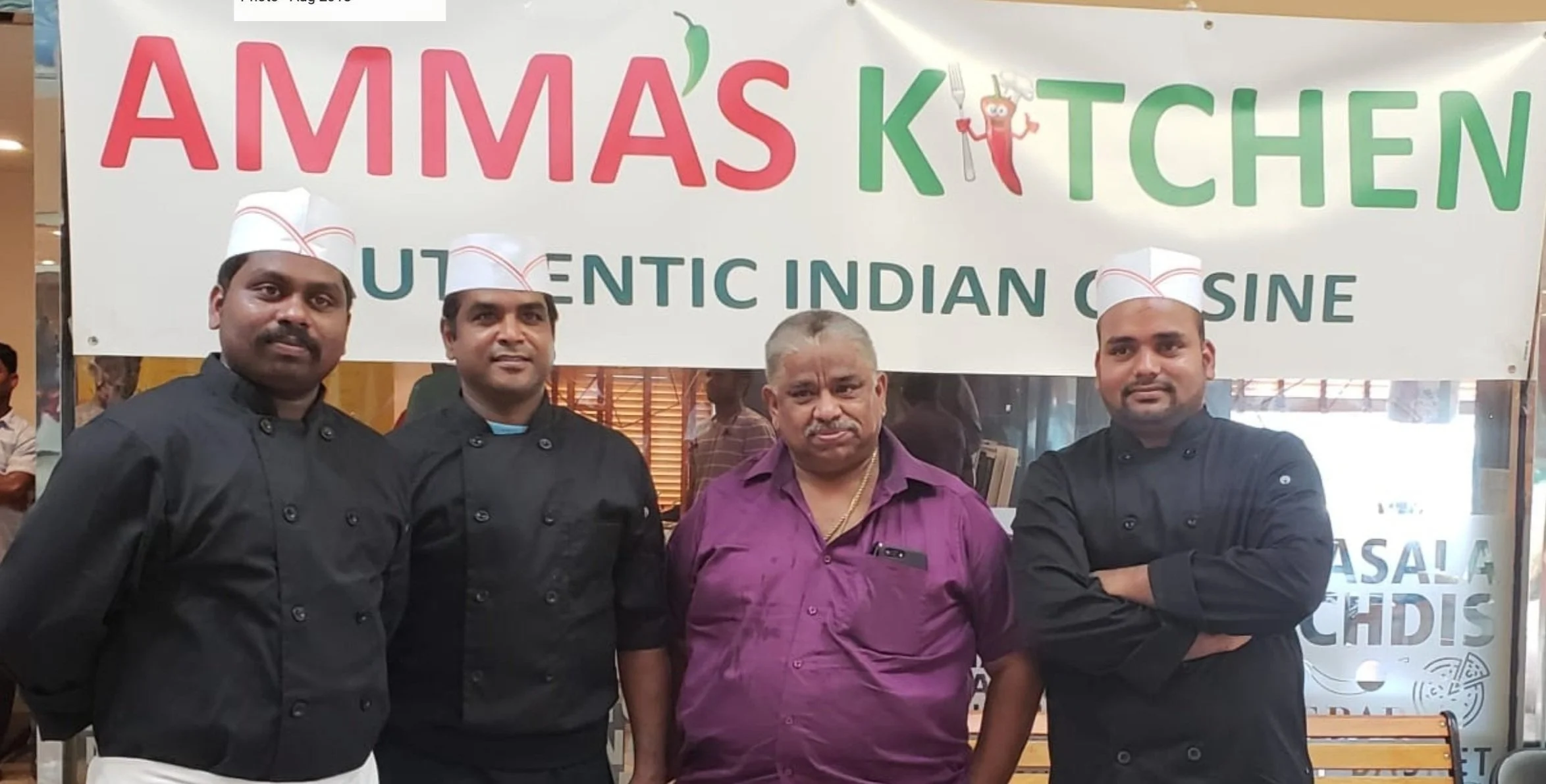Four men standing in front of a restaurant sign that reads 'Amma's Kitchen, Authentic Indian Cuisine.' The two men on the ends are dressed as chefs, wearing black chef jackets and white chef hats. The two middle men are in casual clothing. They are inside a building with a glass window behind them.