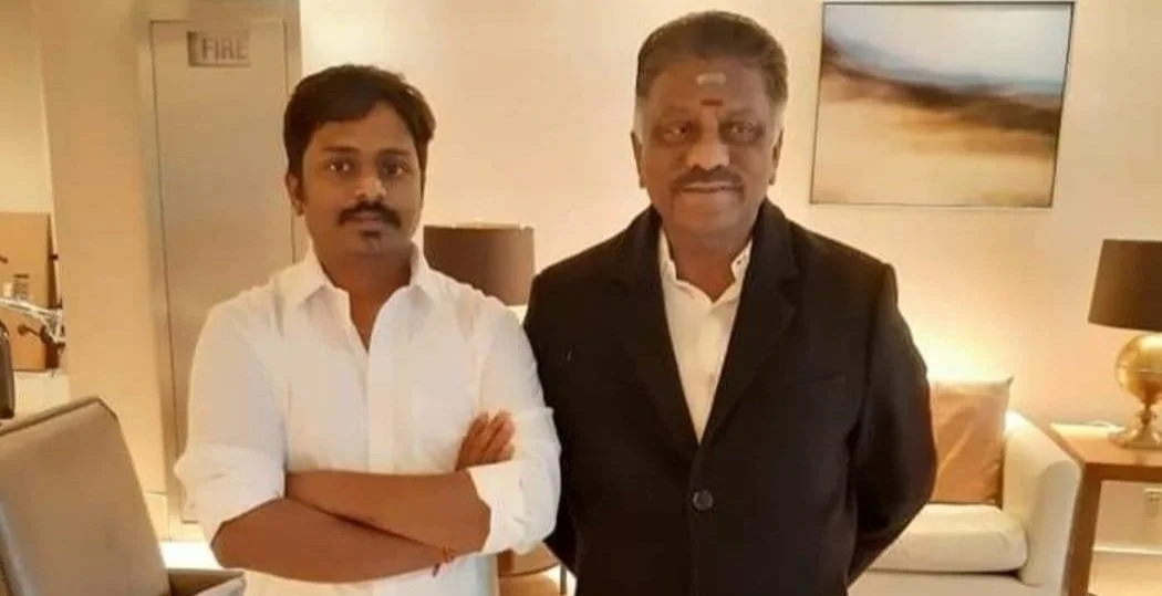 Two men standing side by side in a hotel room, one in a white shirt with arms crossed and the other in a black suit with hands behind his back.